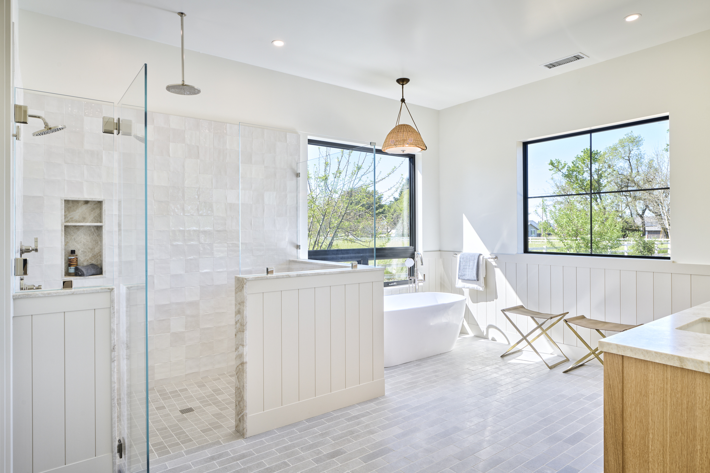 This luxurious primary bathroom features a bright, airy aesthetic with a spacious walk-in shower enclosed by glass, a freestanding soaking tub, and a double vanity. The room is accented by white shiplap wainscoting, light-toned floor tiles, and large windows that flood the space with natural light. A stylish woven pendant light hangs above the tub, creating a serene and spa-like atmosphere.