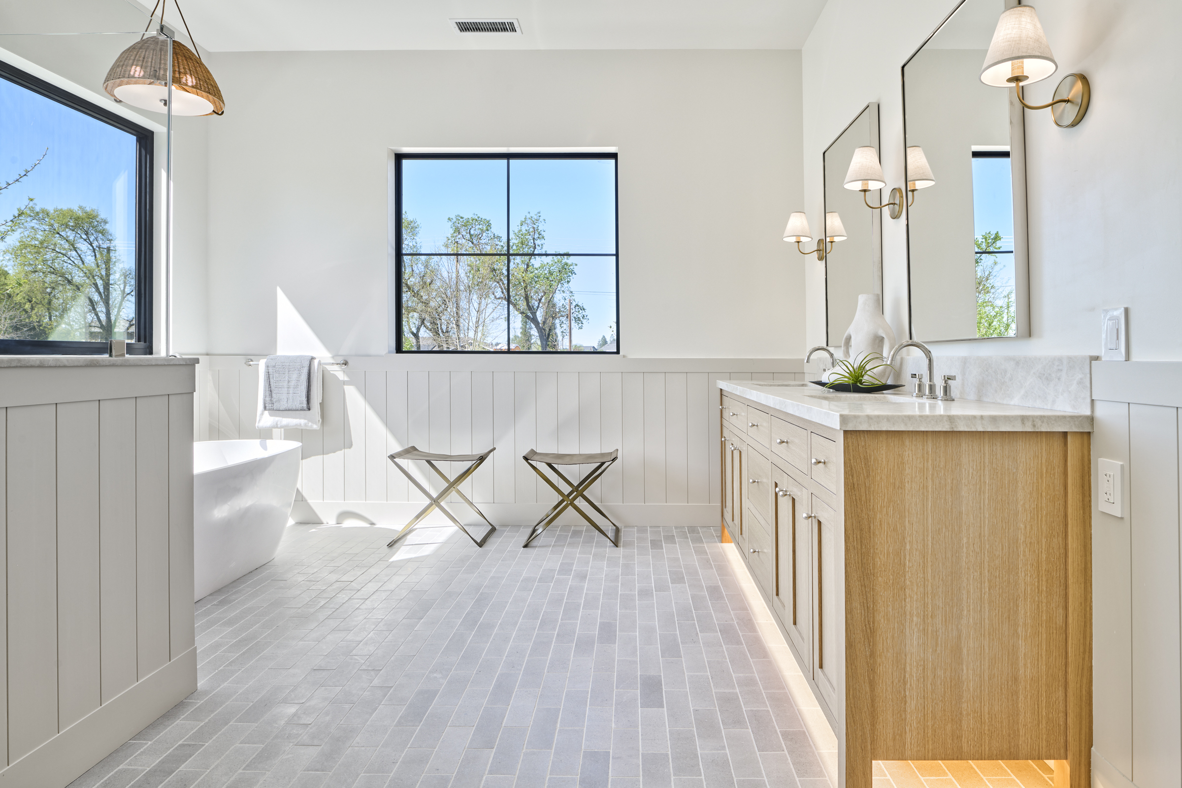 This elegant primary bathroom features a light wood vanity with marble countertops, dual mirrors, and sophisticated wall sconces. A freestanding soaking tub sits beneath a large window, complemented by a clean, minimalist aesthetic with vertical wainscoting and light grey brick-style floor tiles. The space is bright and airy, offering a serene, spa-like atmosphere.