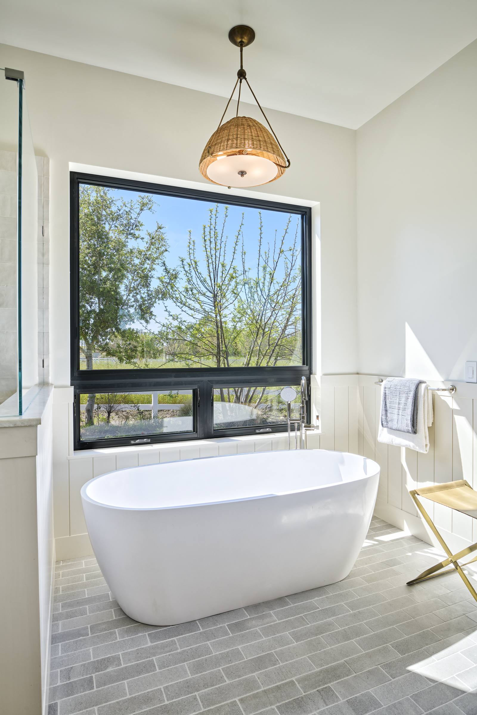 This serene primary bathroom features a sleek, white freestanding soaking tub positioned beneath a large, black-framed window that offers a picturesque view of the outdoors. The space is accented by a textured, woven pendant light fixture and light gray brick-style floor tiles, creating a clean, modern farmhouse aesthetic. A small gold-toned stool and a towel rack add functional elegance to the bright, airy room.