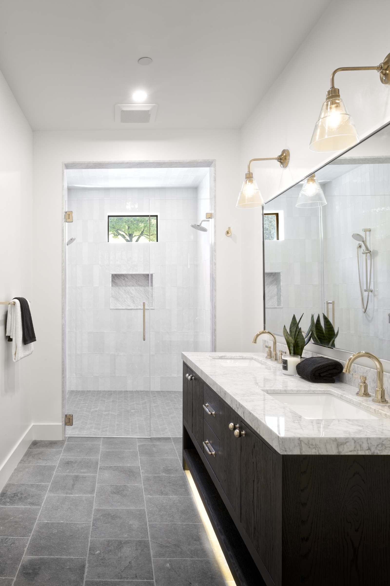 This elegant primary bathroom features a sleek, dark wood double vanity topped with white marble and gold fixtures, creating a sophisticated contrast. A spacious walk-in shower with light-colored tile and a glass door sits at the end of the room, complemented by a large mirror and warm, ambient under-cabinet lighting. The overall aesthetic is modern and luxurious, emphasizing clean lines and high-end finishes.