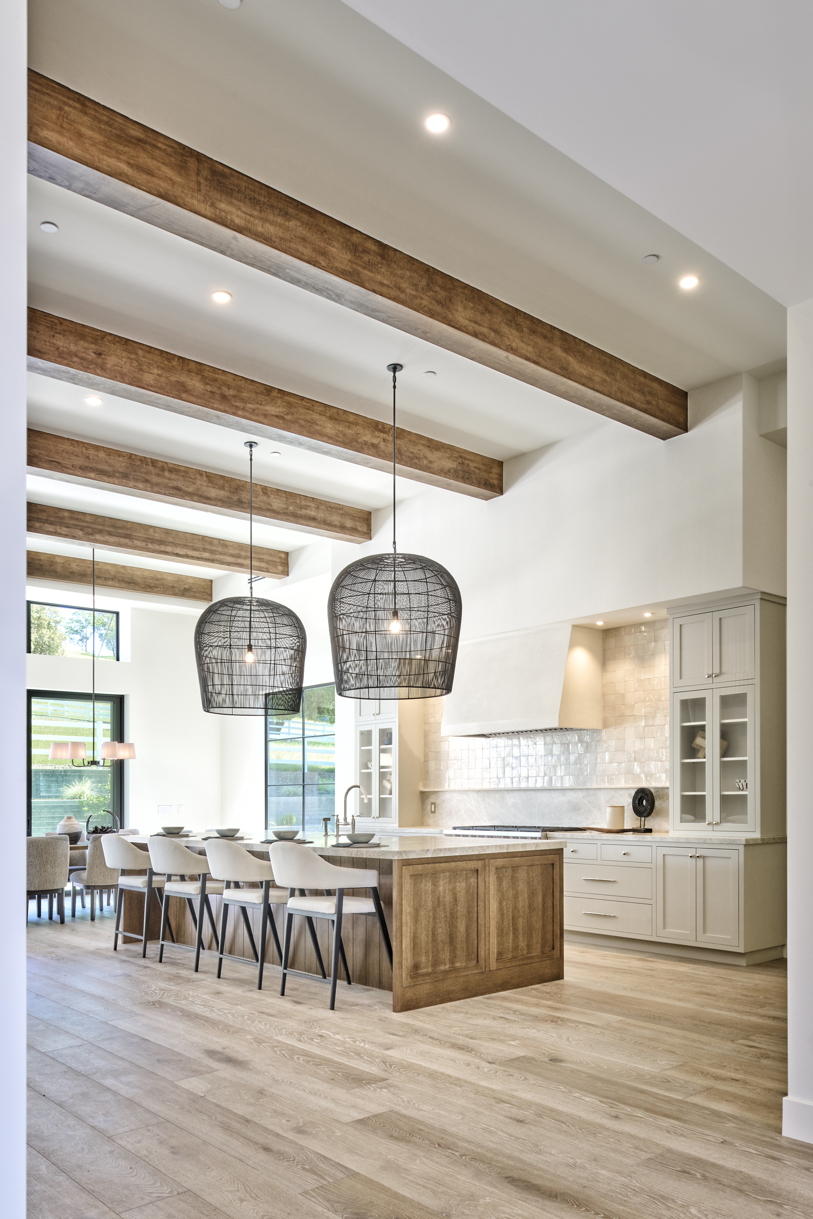 This elegant, high-end kitchen features a large wooden island with bar seating, complemented by light-toned cabinetry and a textured tile backsplash. The space is defined by dramatic exposed wooden ceiling beams and oversized, dark wire-mesh pendant lights that create a sophisticated, airy atmosphere. The perspective is a wide-angle shot from the living area, showcasing the seamless flow into the dining space and the bright, natural light streaming in from the large windows.