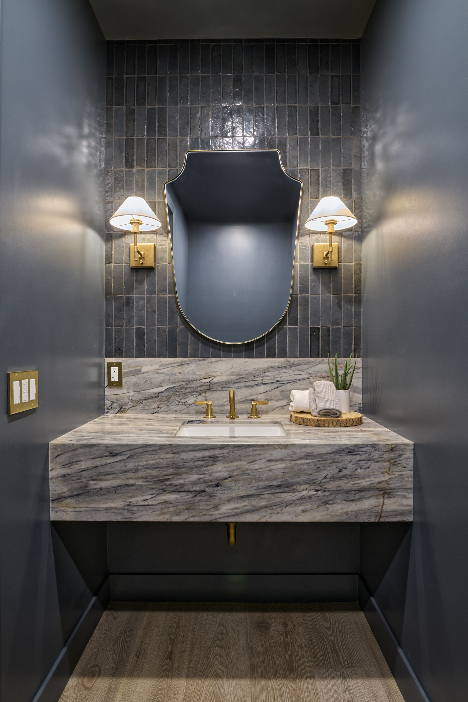 This elegant powder room features a striking floating marble vanity with dramatic veining, complemented by a dark, moody color palette. A unique, shield-shaped mirror is centered against a backdrop of vertical, glossy charcoal tiles, flanked by sophisticated brass wall sconces. The space exudes a high-end, boutique hotel aesthetic, perfect for a luxury home listing.