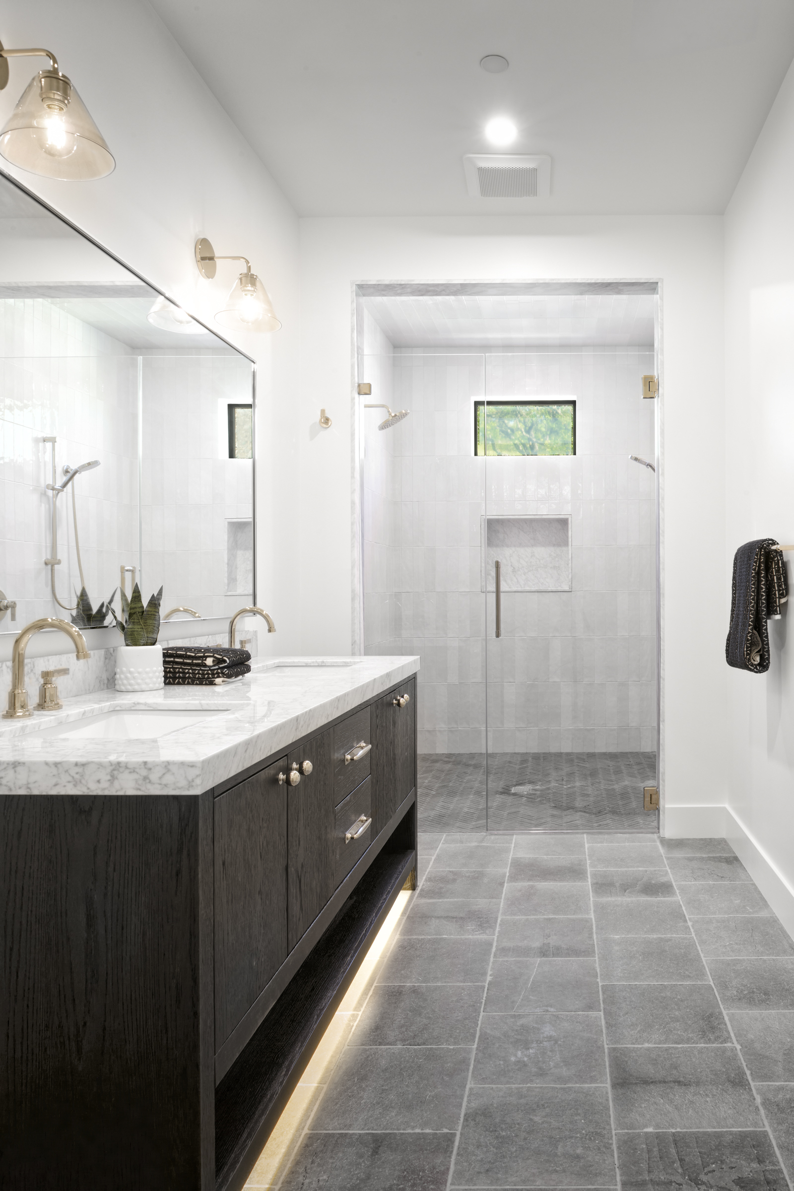 This modern primary bathroom features a sleek dark wood vanity with a marble countertop, dual gold-toned faucets, and under-cabinet accent lighting. A spacious walk-in shower with glass doors and a small window is positioned at the back, complemented by grey stone floor tiles. The overall aesthetic is sophisticated and contemporary, emphasizing clean lines and high-end finishes.