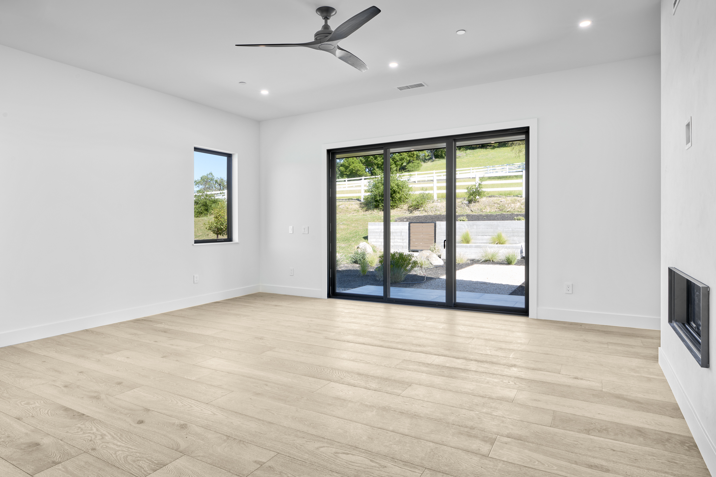 This bright and airy living room features light-toned wood flooring, crisp white walls, and a modern ceiling fan, creating a clean and minimalist aesthetic. A large sliding glass door provides a seamless transition to the outdoor patio and landscaped backyard, filling the space with natural light. The room is anchored by a sleek, built-in fireplace on the right, offering a contemporary focal point for the open-concept living area.