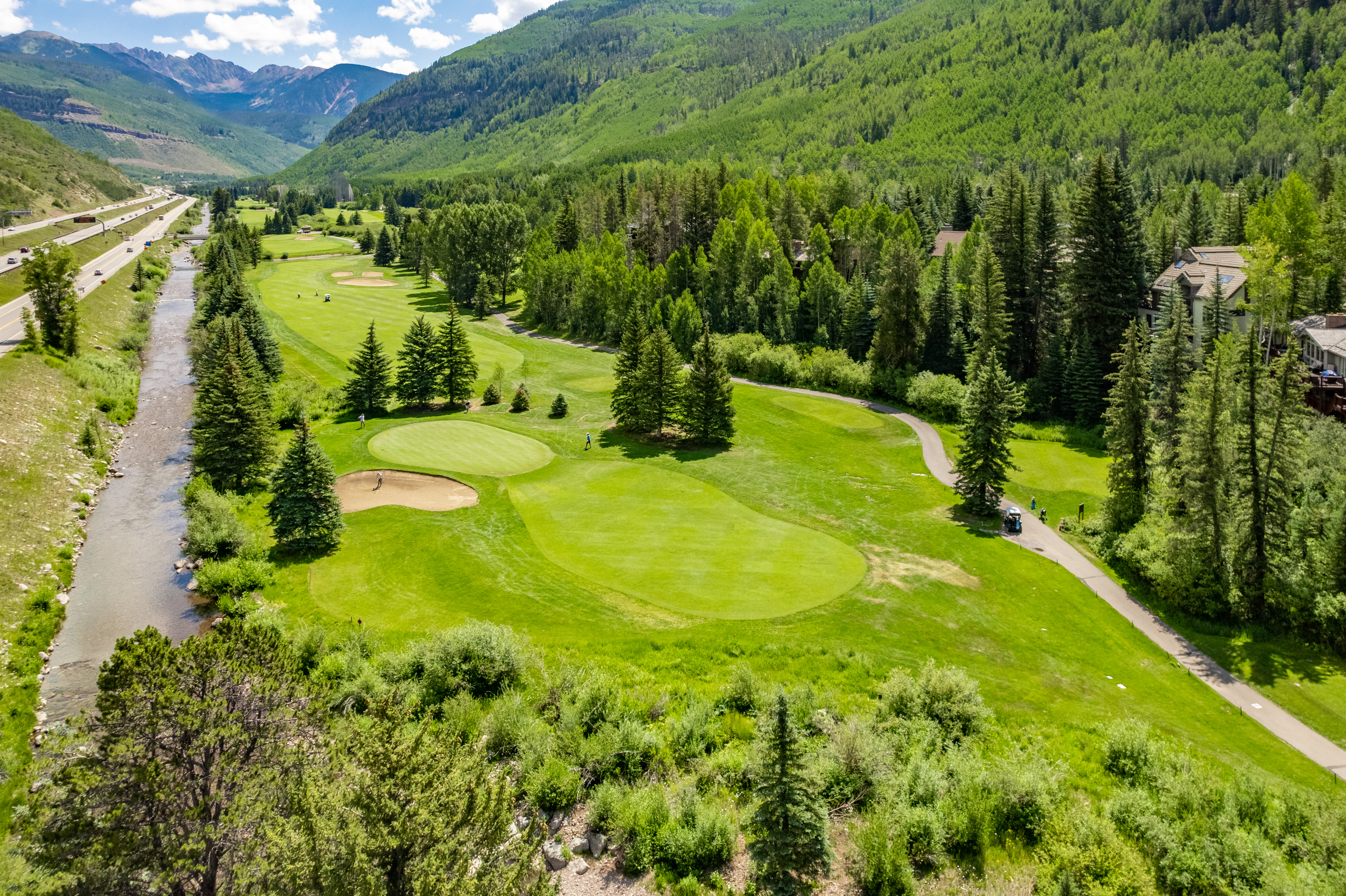 This stunning aerial view captures a scenic golf course nestled within a lush, mountainous landscape alongside a meandering river and a nearby highway. The property features well-manicured greens, dense forest integration, and provides direct access to convenient paved cart paths for leisure. The idyllic setting highlights the balance between outdoor recreational living and natural beauty, making it a compelling feature for potential homeowners.