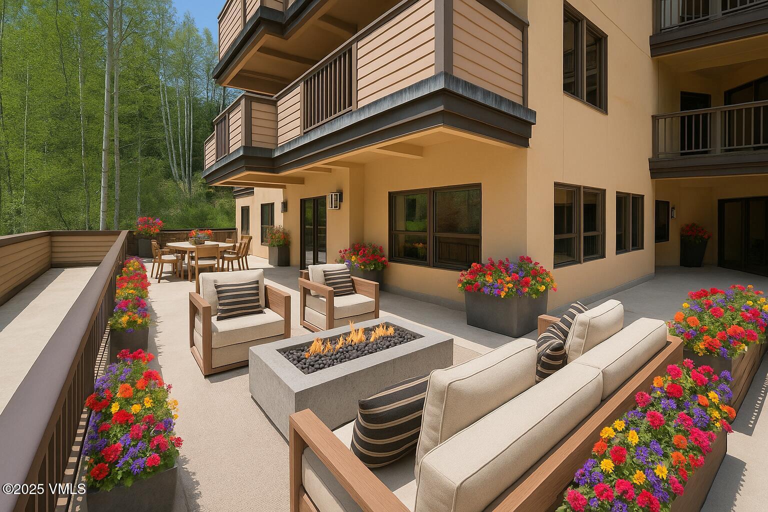 This spacious, modern patio offers an inviting outdoor living area, beautifully landscaped with vibrant floral planters. The space is equipped with comfortable outdoor seating, a central rectangular gas fire pit, and a separate dining area, making it perfect for entertaining. The setting is enhanced by scenic, lush green mountain views in the backdrop, reflecting a high-end, relaxed lifestyle.