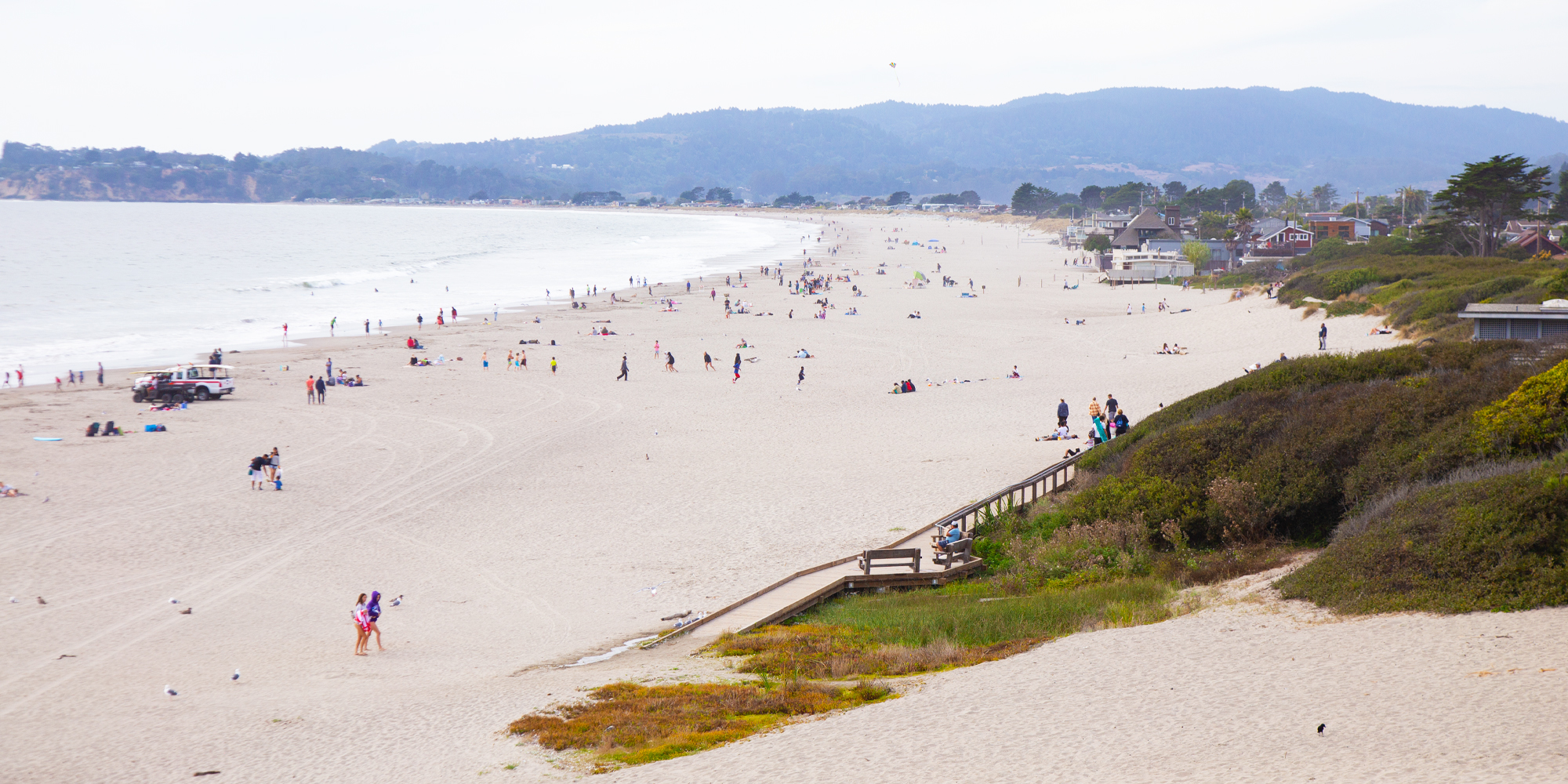 This panoramic outdoor shot captures a vast, sandy public beach featuring a wooden boardwalk accessible from a vegetated dune area. With rolling hills in the background and numerous visitors enjoying the coastline, the scene highlights the beach as a prominent community amenity. The open space and natural coastal setting provide an inviting atmosphere for potential local residents.