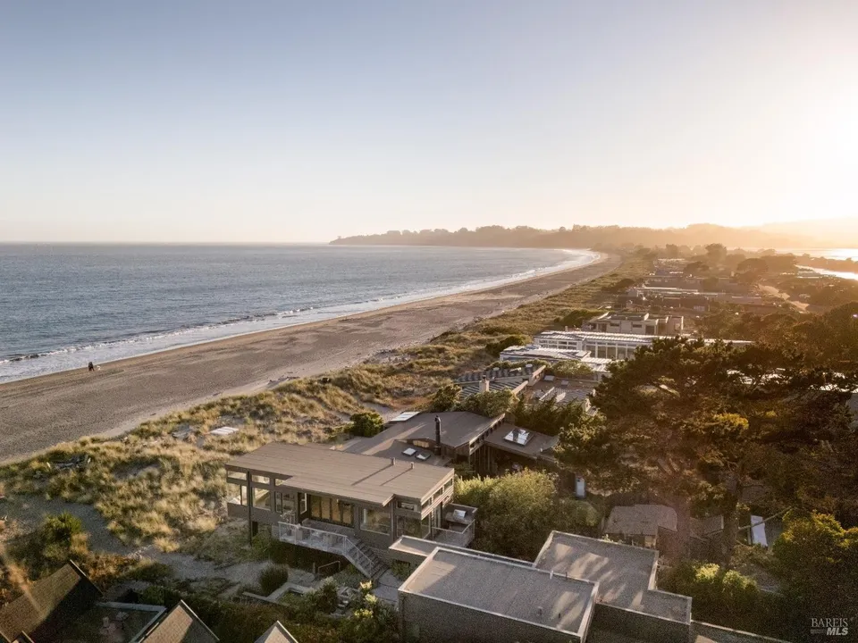 This is a scenic, high-angle aerial photograph of a beachfront property featuring contemporary architectural design with flat roofs and multiple levels. The house sits nestled amongst coastal dunes and established trees, offering unparalleled ocean views and direct beach access. The serene setting and modern structural profile suggest a high-end coastal retreat.