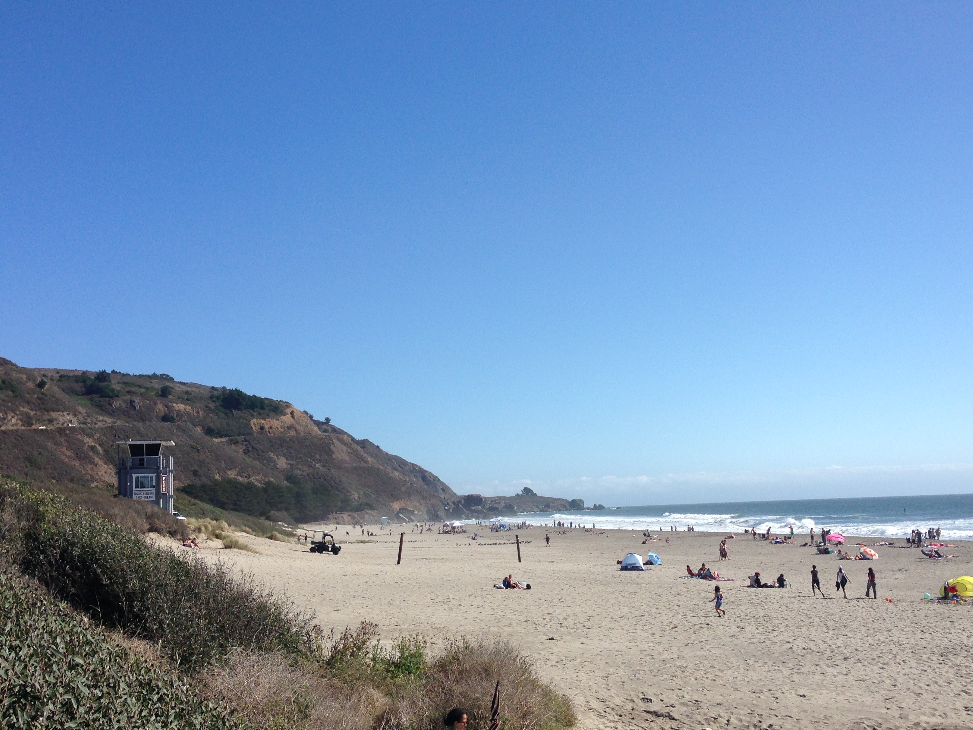 This image features a scenic public beach setting with a prominent lifeguard tower overlooking the sandy shoreline and ocean. The expansive landscape includes a hillside backdrop and open space suitable for outdoor recreation, making it an attractive recreational amenity for nearby residents and visitors.