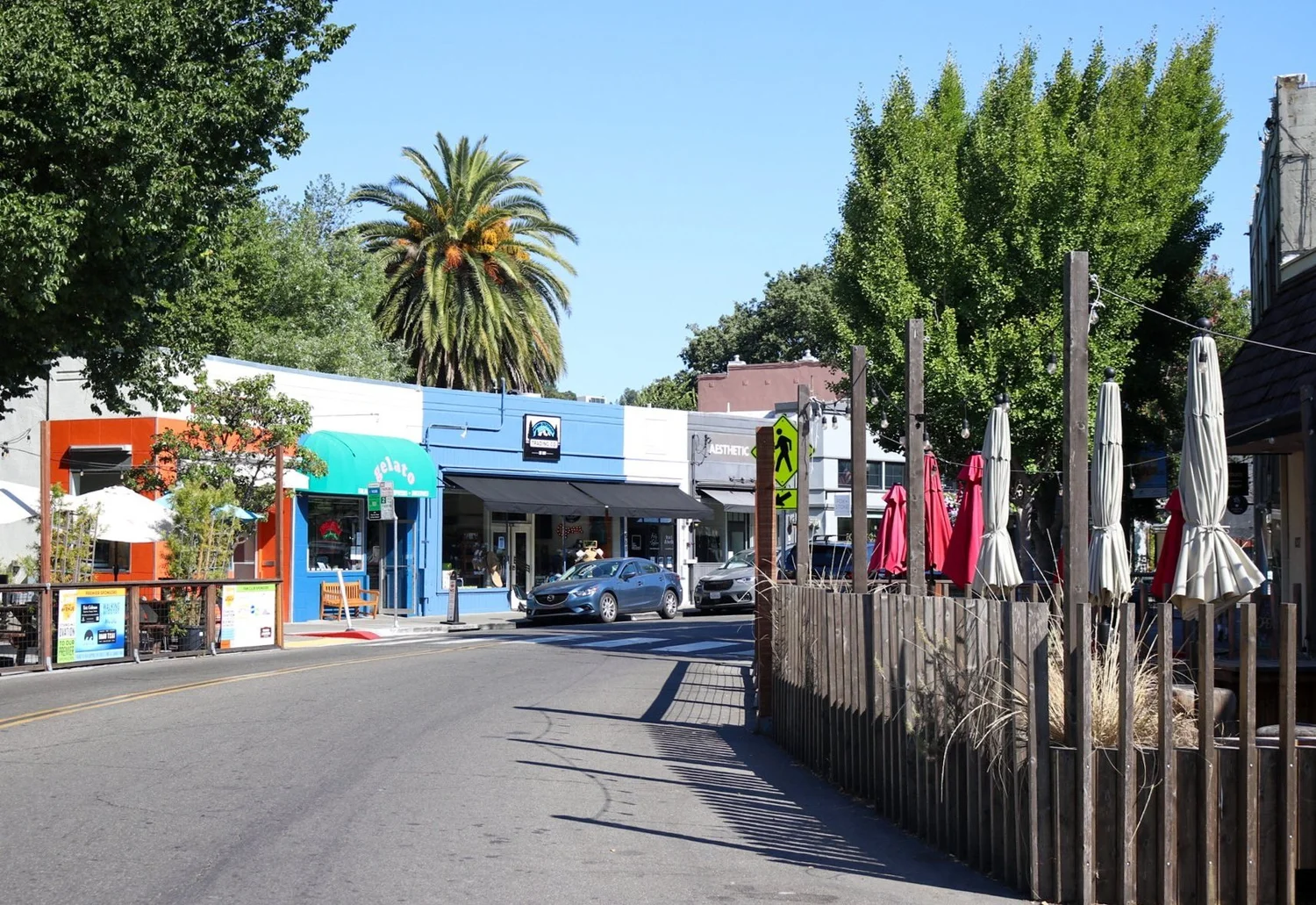 This image displays a vibrant commercial street scene featuring a row of small, distinctive storefronts with a variety of architectural colors and styles. A pedestrian-friendly area with outdoor patio seating protected by a wooden fence, colorful umbrellas, and a prominent palm tree creates a welcoming neighborhood atmosphere. The sunny environment highlights the walkability and charm of this local business district.