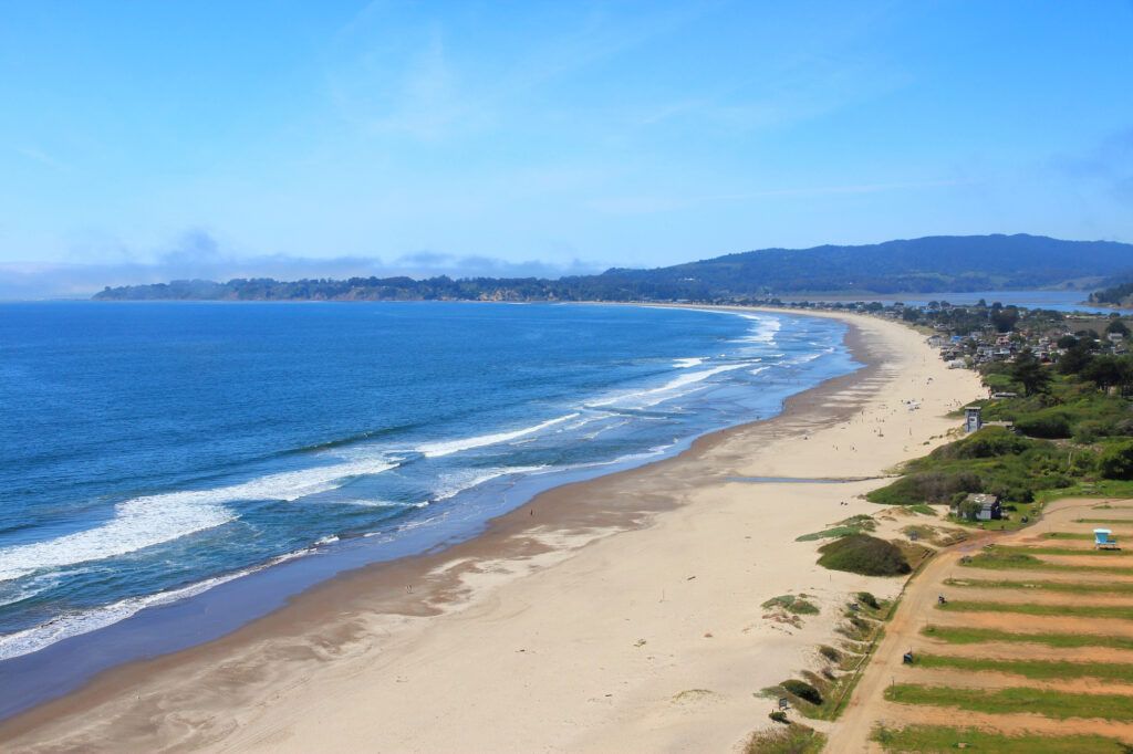 This stunning aerial view captures a picturesque coastline with a vast sandy beach stretching towards distant hills. The landscape features a pristine shoreline meeting the blue ocean, complemented by a rustic dirt path and natural vegetation along the dunes. The setting offers an exceptional sense of openness and tranquility, highlighting the prime coastal location of the surrounding area.