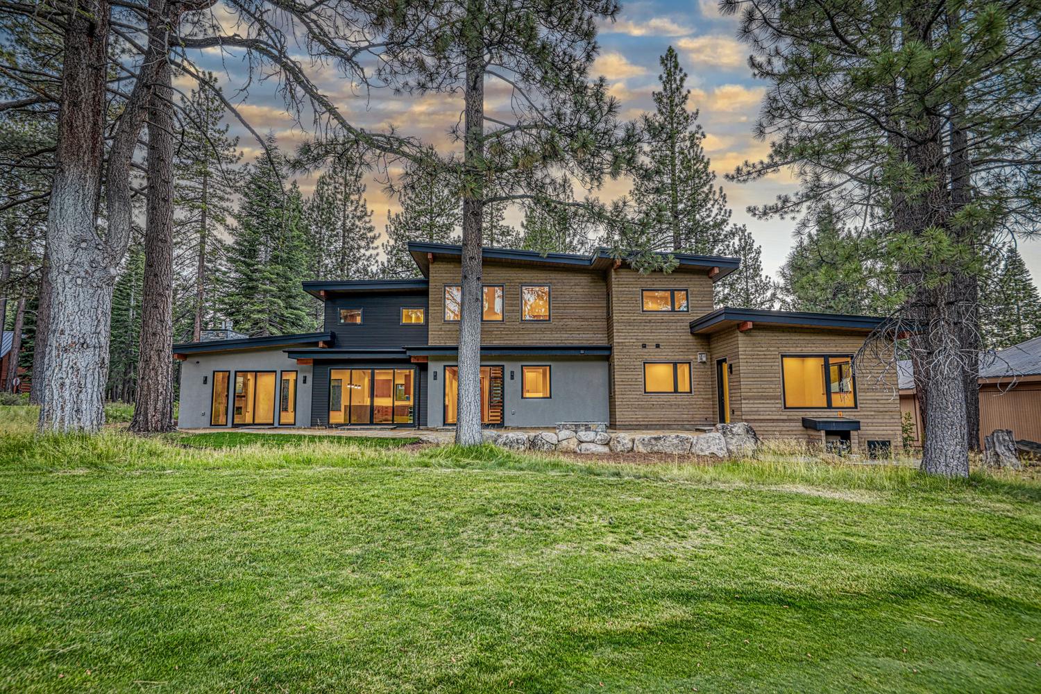 This contemporary mountain home features a striking mix of horizontal wood siding and dark metal accents, set against a backdrop of tall pine trees. The rear elevation showcases large windows and sliding glass doors that open onto a stone patio, creating a seamless connection between the indoor living space and the expansive grassy yard. The architectural design emphasizes clean lines and a modern aesthetic, perfectly integrated into the natural, wooded landscape.