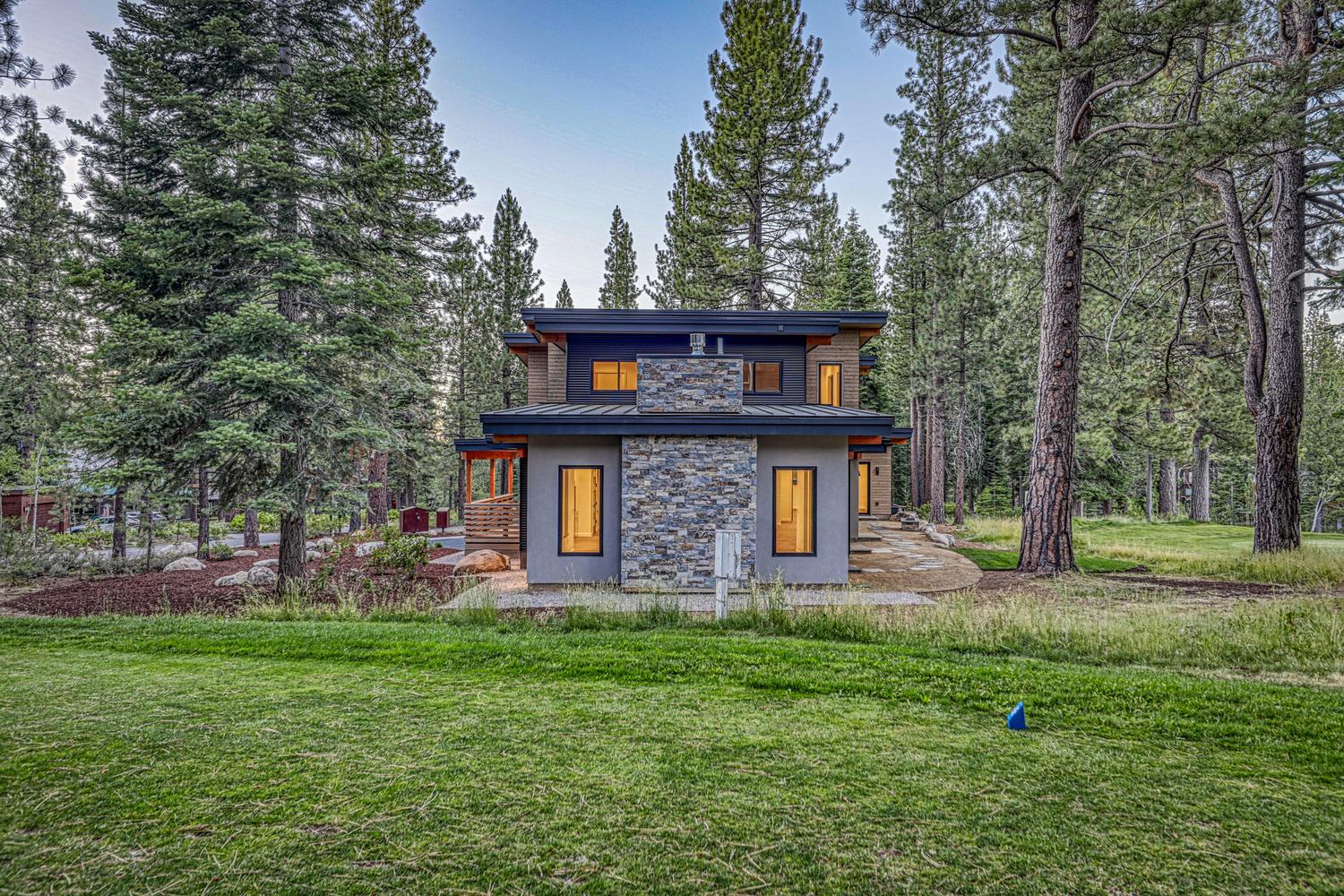 This modern mountain home features a striking combination of gray stucco, horizontal wood siding, and a prominent stone-clad chimney breast centered on the rear facade. The structure is nestled within a lush forest of tall pine trees, with a manicured lawn in the foreground and a stone pathway leading along the side of the house. The design emphasizes clean lines and a contemporary aesthetic, perfectly integrated into its natural, wooded surroundings.