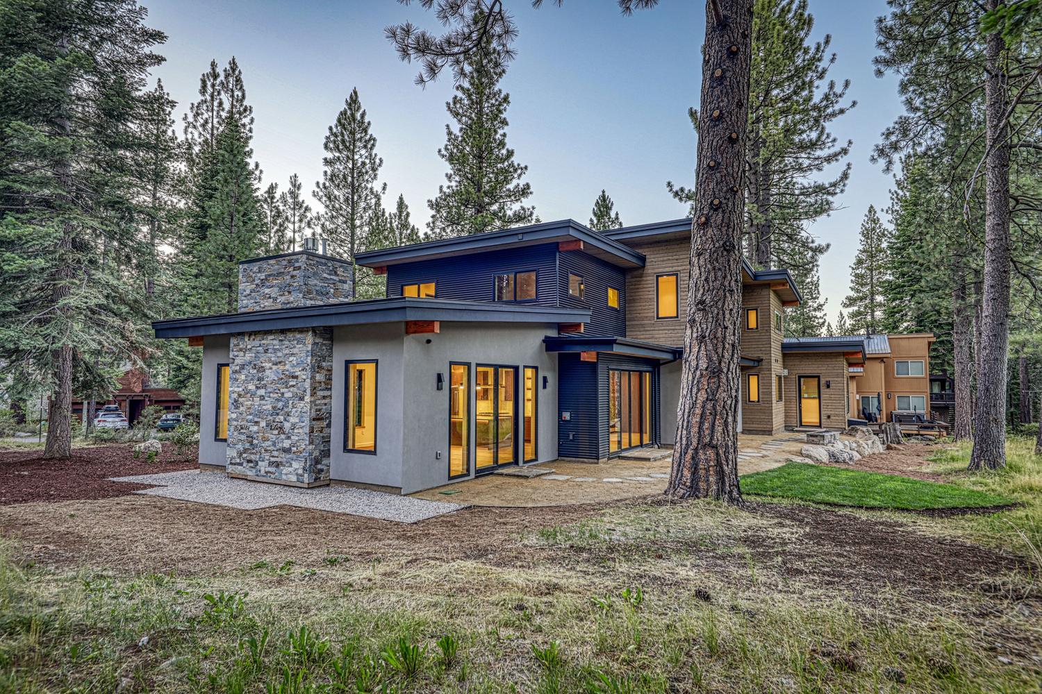 This modern mountain residence features a striking mix of materials, including a prominent stone-clad chimney breast, dark horizontal siding, and warm-toned wood accents. The home is nestled within a dense pine forest, with large glass doors and windows that create a seamless connection to the surrounding natural landscape. A stone patio extends from the rear of the house, providing a serene outdoor space that blends into the wooded environment.