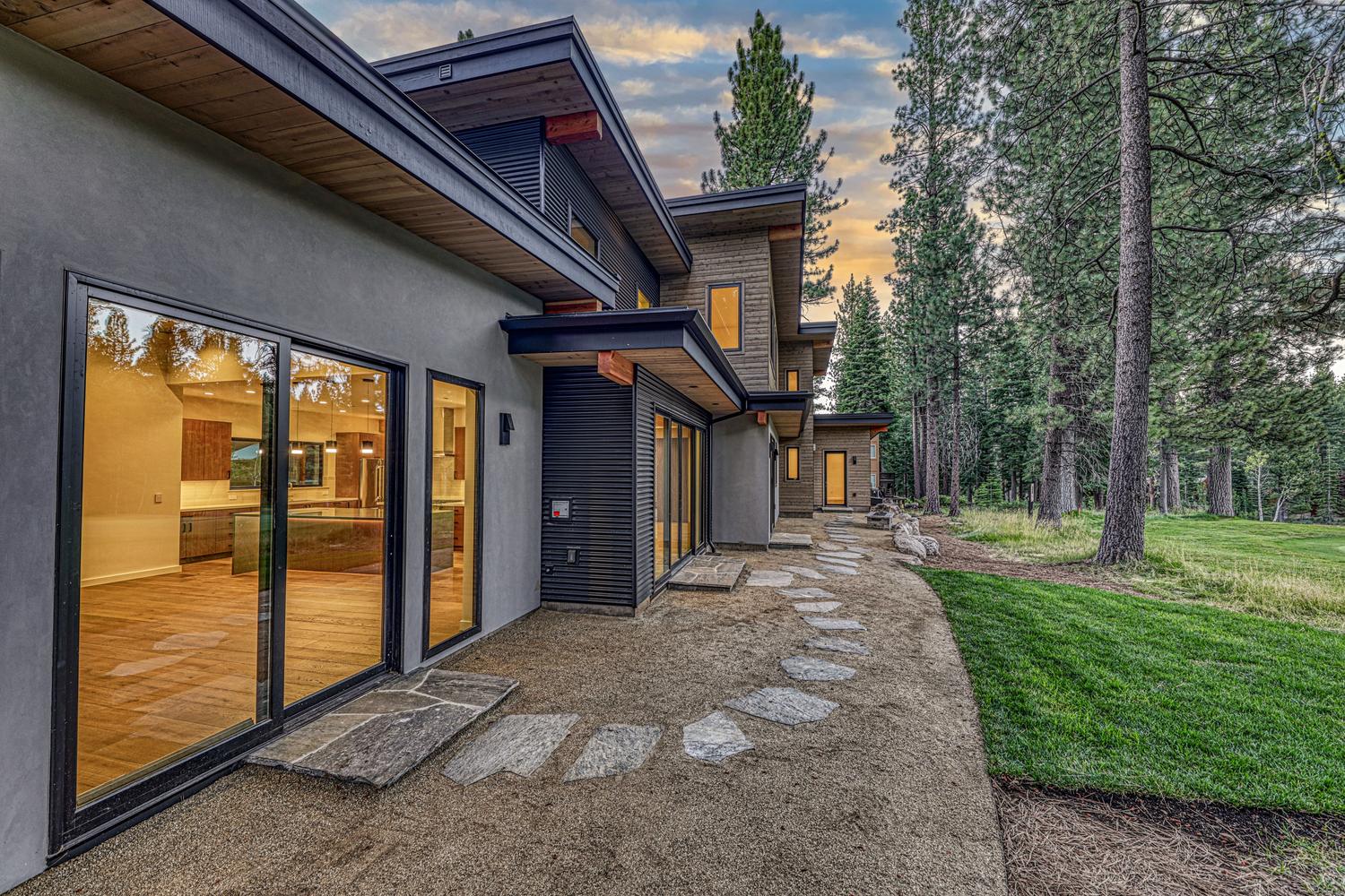 This modern home features a striking exterior with a mix of smooth gray stucco and dark horizontal siding, accented by natural wood soffits. A stone-paved path winds through a gravel area alongside large glass sliding doors that provide a seamless view into the interior living space. The scene is set against a backdrop of tall pine trees under a soft, dusk-lit sky, creating a serene and sophisticated atmosphere.