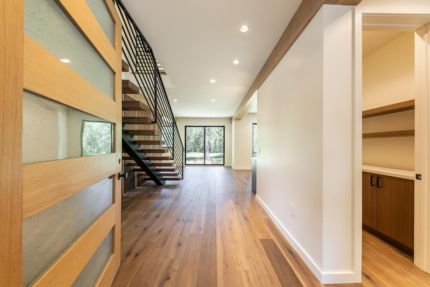 This bright, modern entryway features a striking staircase with open-tread wooden steps and a sleek black metal railing. The perspective looks down a wide hallway with light wood flooring, leading toward a large glass door that provides a clear view of the outdoors. To the right, a doorway opens into a small, organized storage area with built-in shelving and cabinetry, emphasizing the home's clean, contemporary design.