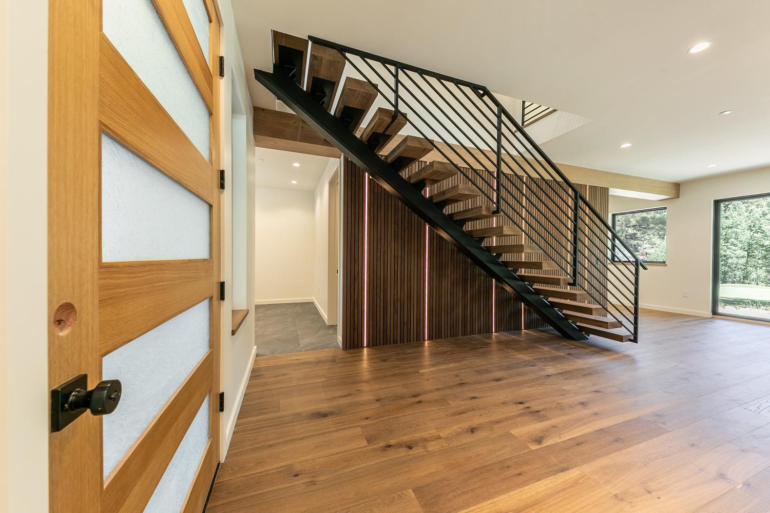 This modern entryway features a striking floating staircase with wood treads and a black metal railing, serving as the focal point of the space. The area is accented by a wall with vertical wood slats and integrated vertical lighting, creating a warm and sophisticated atmosphere. Wide-plank wood flooring flows throughout the open-concept layout, which includes a view into a secondary hallway and large windows that bring in natural light.