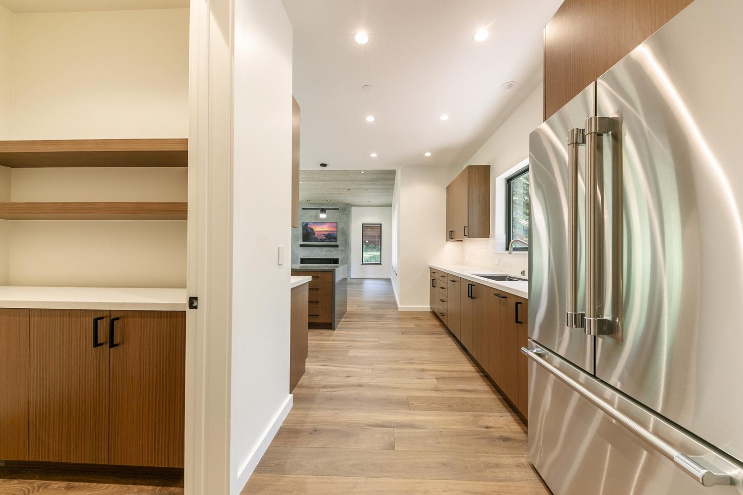 This modern kitchen features warm wood cabinetry paired with light-toned countertops and stainless steel appliances, including a prominent refrigerator in the foreground. The perspective looks through a transition space into a bright, open-concept living area with a television mounted above a fireplace. The design emphasizes clean lines, natural wood flooring, and a seamless flow between the functional kitchen workspace and the adjacent living room.