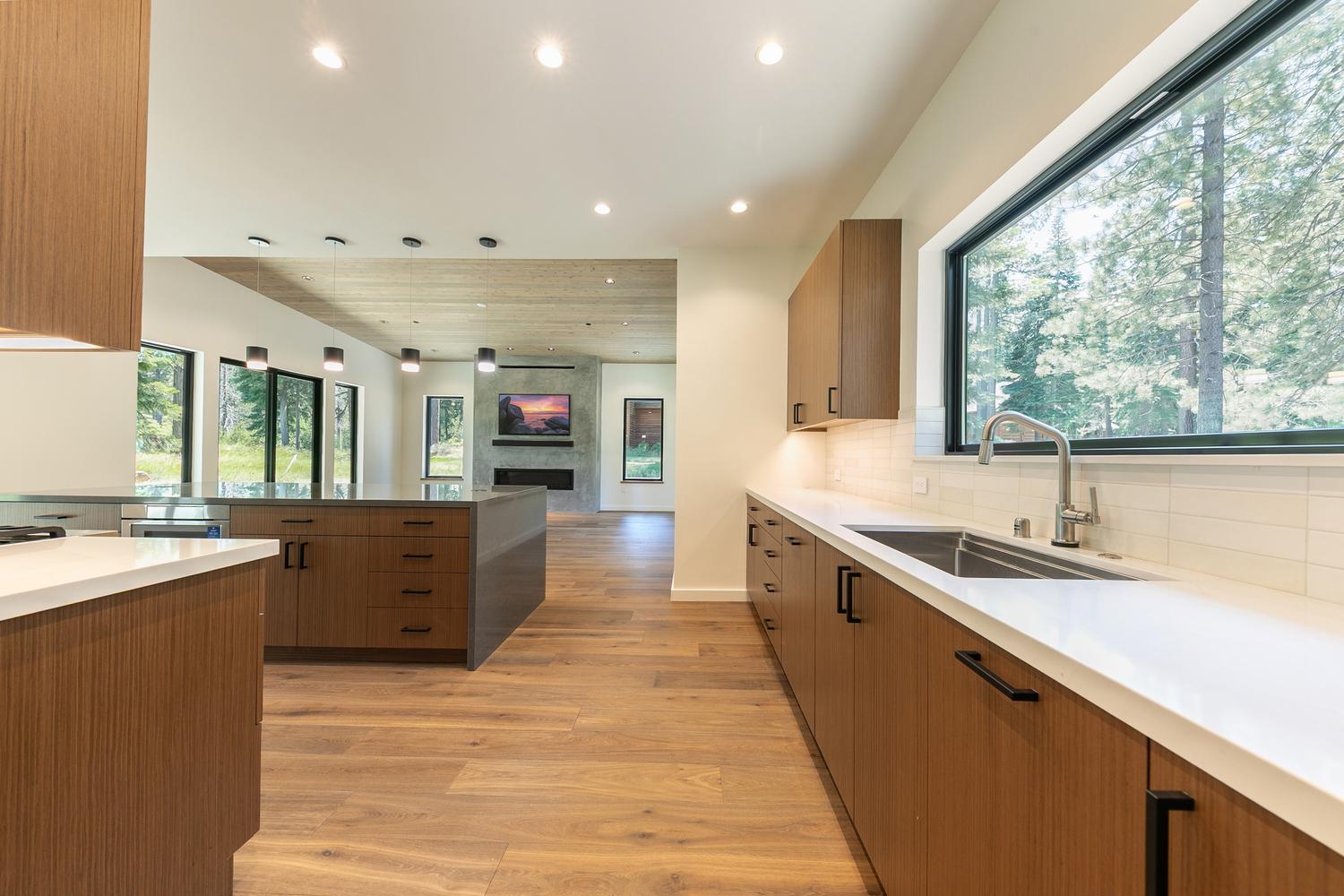 This modern kitchen features warm wood-toned cabinetry paired with sleek white countertops and a long, central island. A large window above the sink provides a bright, natural view of the surrounding trees, while the open-concept layout flows seamlessly into a living area with a stone-clad chimney breast. The space is illuminated by recessed lighting and minimalist pendant lights, creating a clean, contemporary aesthetic.