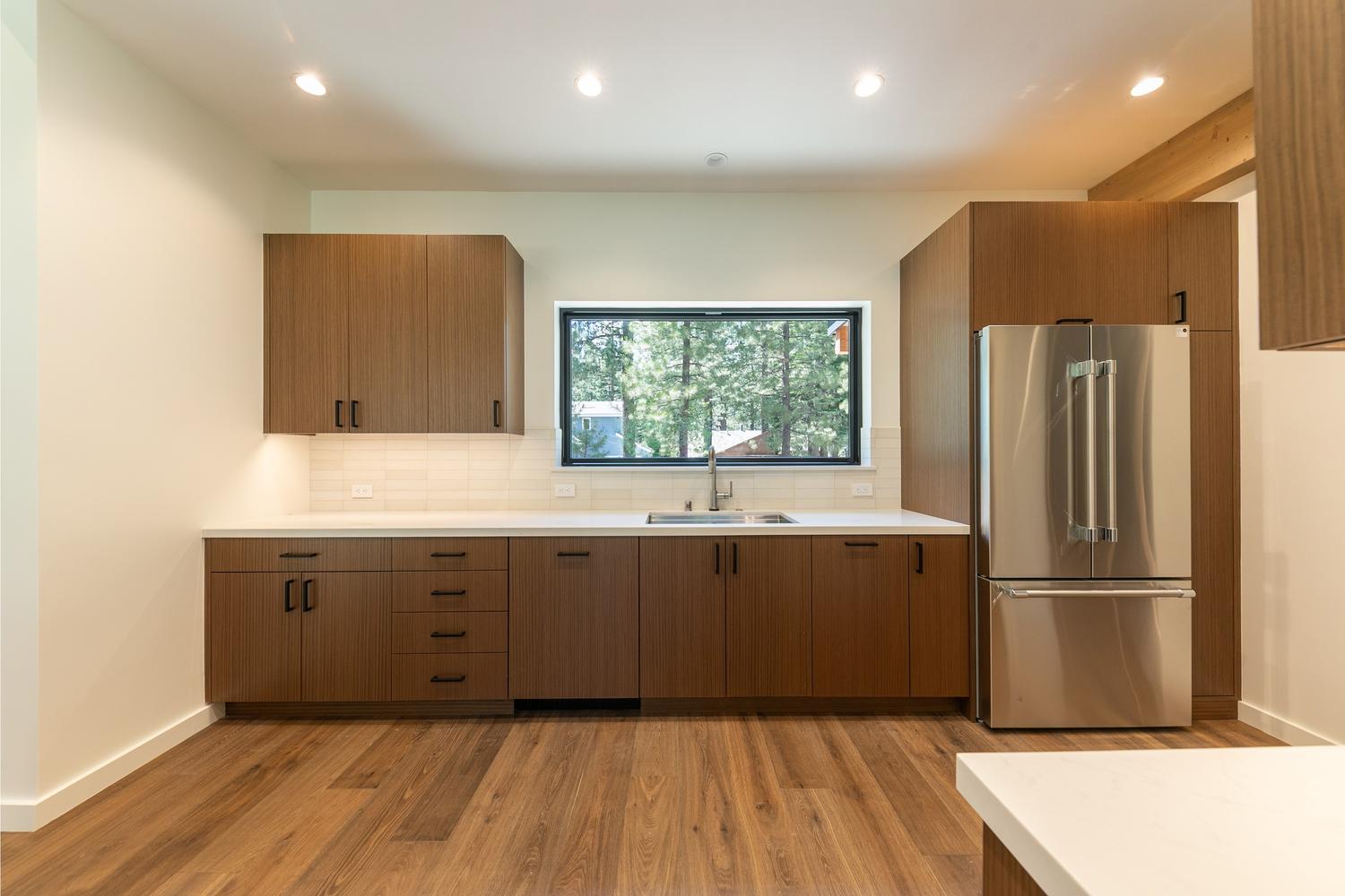This modern kitchen features sleek, dark wood-toned cabinetry paired with white countertops and a light-colored tiled backsplash. A stainless steel refrigerator is integrated into the cabinetry, and a central window provides a clear view of the surrounding trees. The perspective is a straight-on, eye-level shot that highlights the clean lines and contemporary aesthetic of the space.