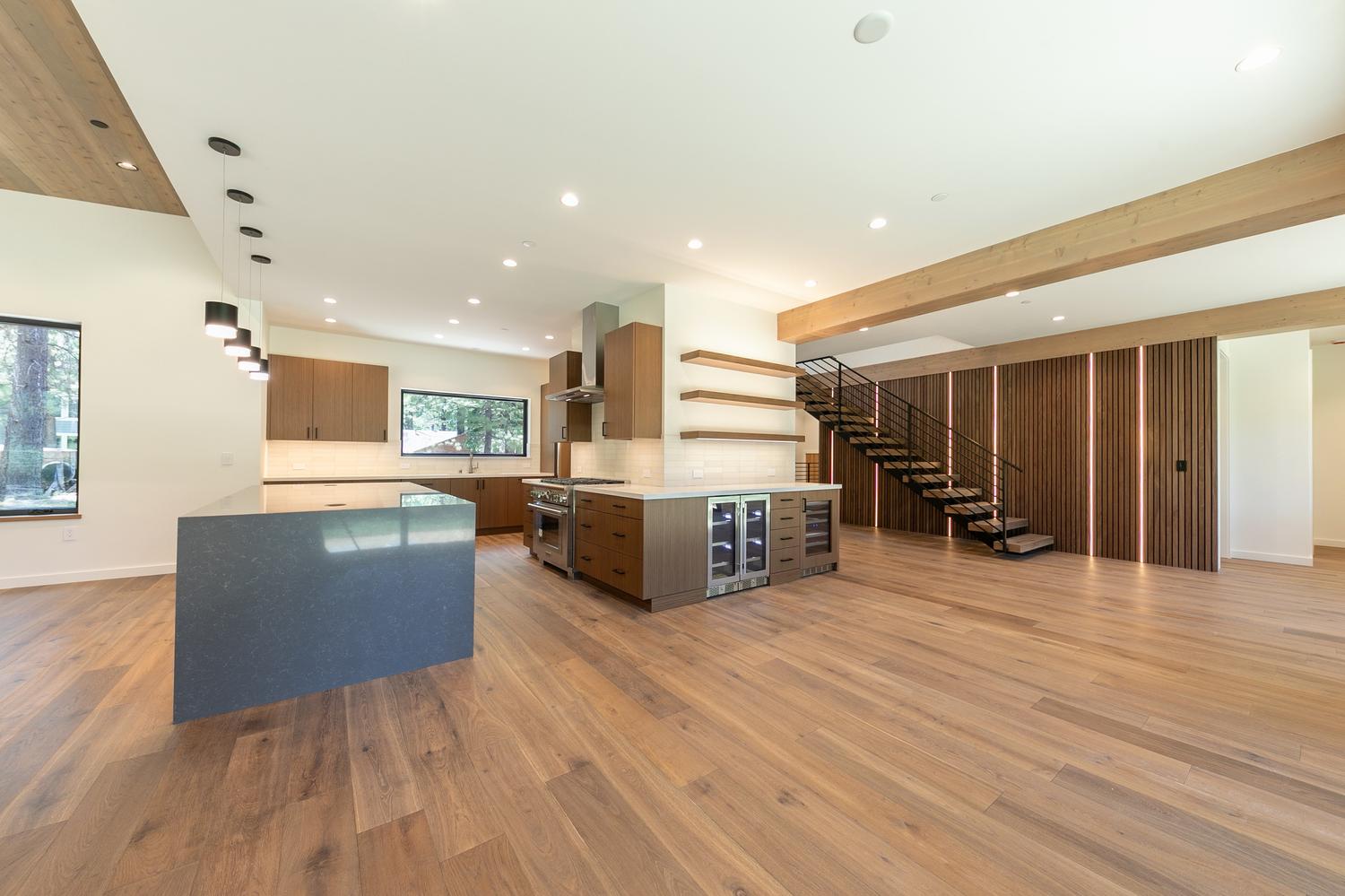 This open-concept kitchen features a large central island with a dark stone countertop and warm wood cabinetry. The space is anchored by a professional-grade range, stainless steel appliances, and floating shelves, all set against a backdrop of light-toned walls and wide-plank wood flooring. To the right, a staircase with a modern metal railing leads upward, adjacent to a decorative wood-slat wall accented with integrated vertical lighting.