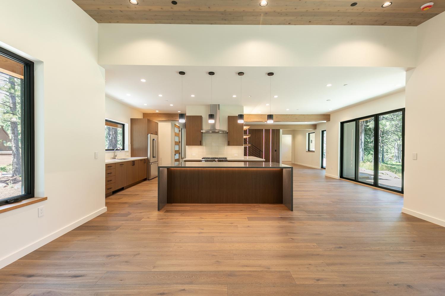 This spacious, open-concept kitchen features a large central island with a dark base and a light-toned countertop, complemented by warm wood cabinetry and stainless steel appliances. The room is illuminated by a series of modern pendant lights and recessed lighting, with large windows and glass doors providing ample natural light and views of the surrounding trees. The design aesthetic is clean and contemporary, characterized by wide-plank wood flooring and a neutral color palette that creates an inviting, airy atmosphere.