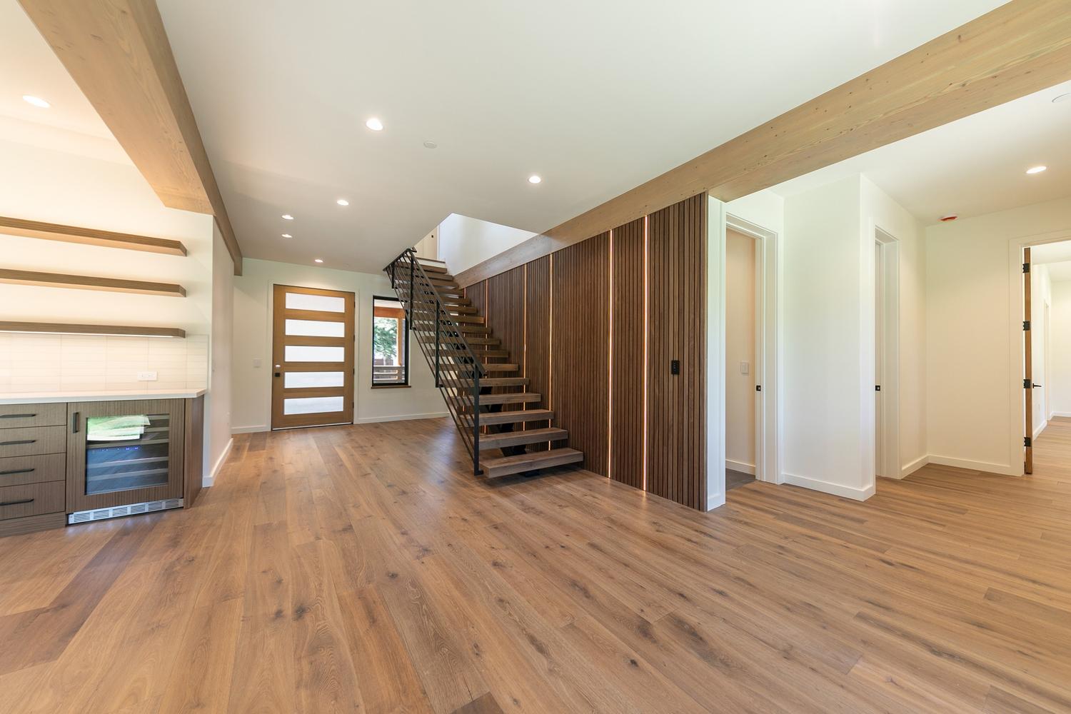This bright and modern entryway features a striking staircase with open-riser wood treads and a sleek black metal railing. To the right, a wall of vertical wood slats with integrated linear lighting creates a warm, textured focal point, while the left side includes a built-in beverage cooler and floating shelves. The open-concept layout flows seamlessly across light-toned wood flooring, leading toward multiple doorways and a front door with horizontal glass panels.