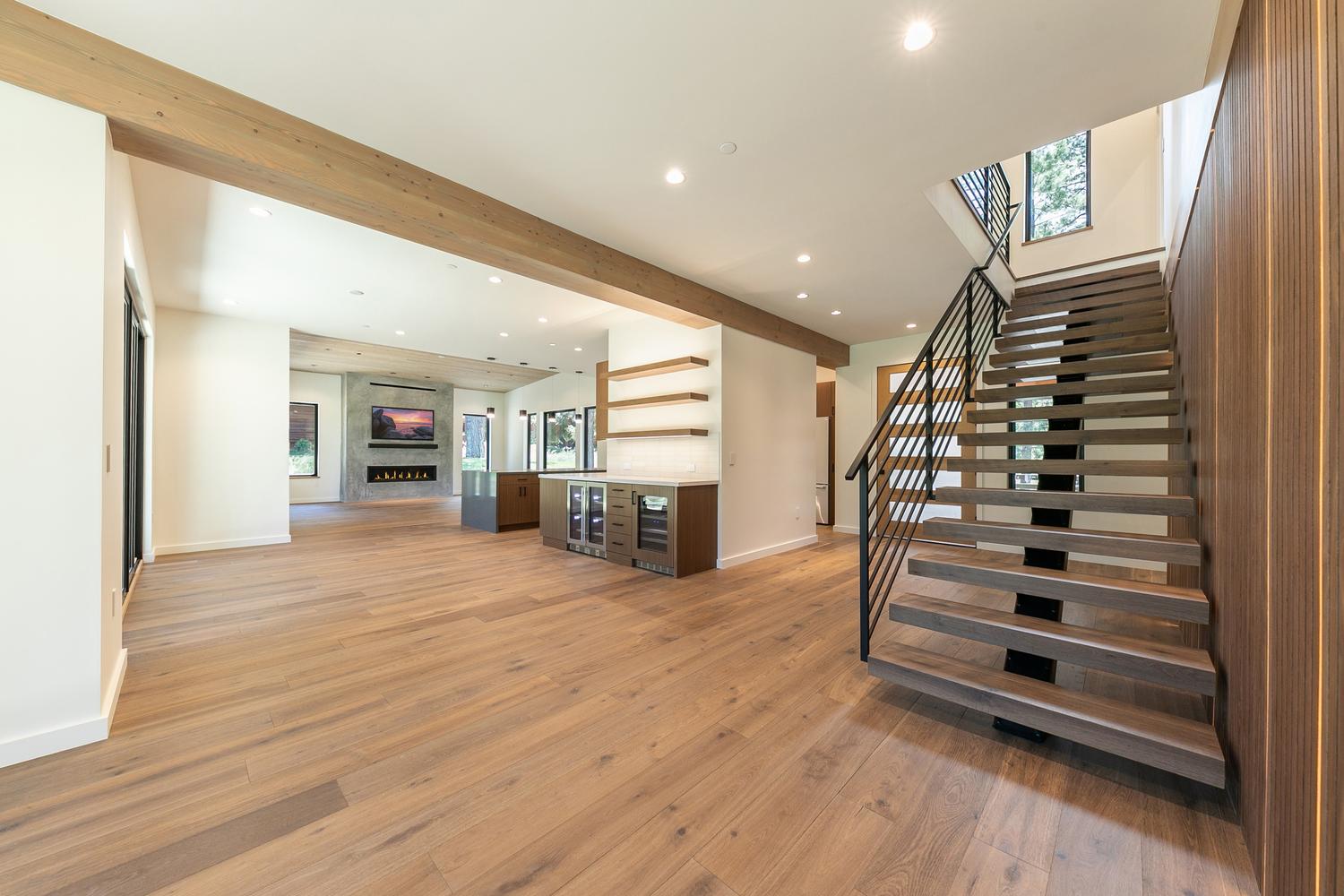 This open-concept interior space features a striking floating staircase with dark metal railings and wood treads, serving as a central architectural focal point. The area flows seamlessly into a kitchen and living space, characterized by light wood flooring, a tall stone-clad chimney breast, and exposed wood ceiling beams. The perspective captures the airy, modern design, emphasizing the clean lines and the transition between the entry staircase and the main living area.