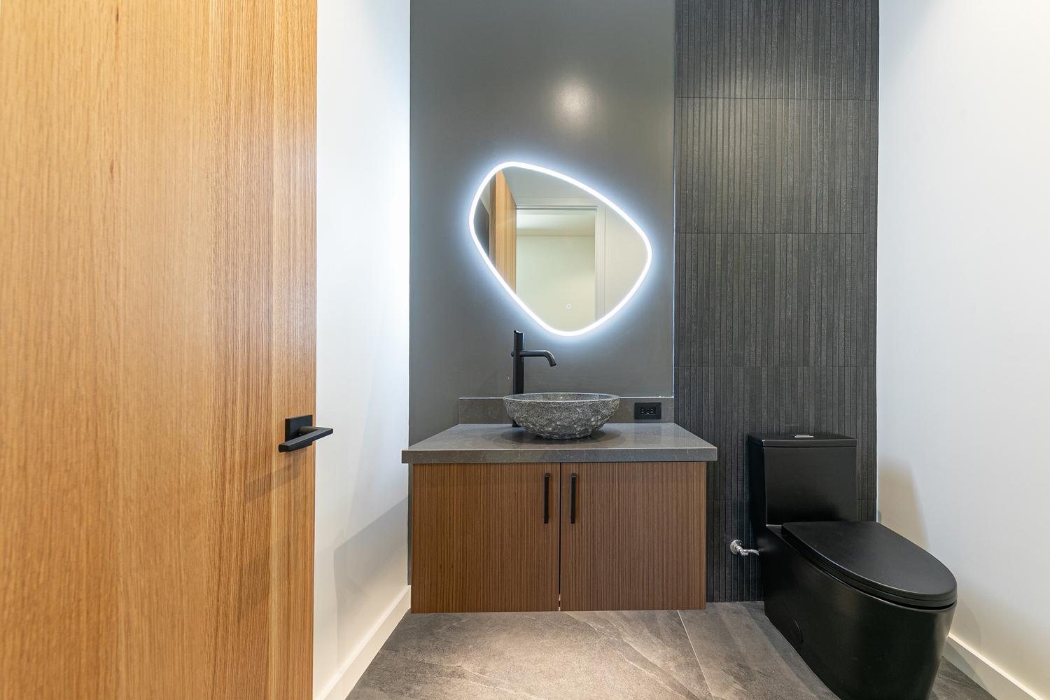 This modern powder room features a floating wood vanity topped with a stone counter and a unique, textured stone vessel sink. A striking, organic-shaped LED-backlit mirror serves as a focal point against a dark, smooth wall, while the adjacent wall is clad in vertical, dark-toned textured tiles. The space is completed by a sleek black toilet and large-format grey floor tiles, creating a sophisticated and contemporary aesthetic.