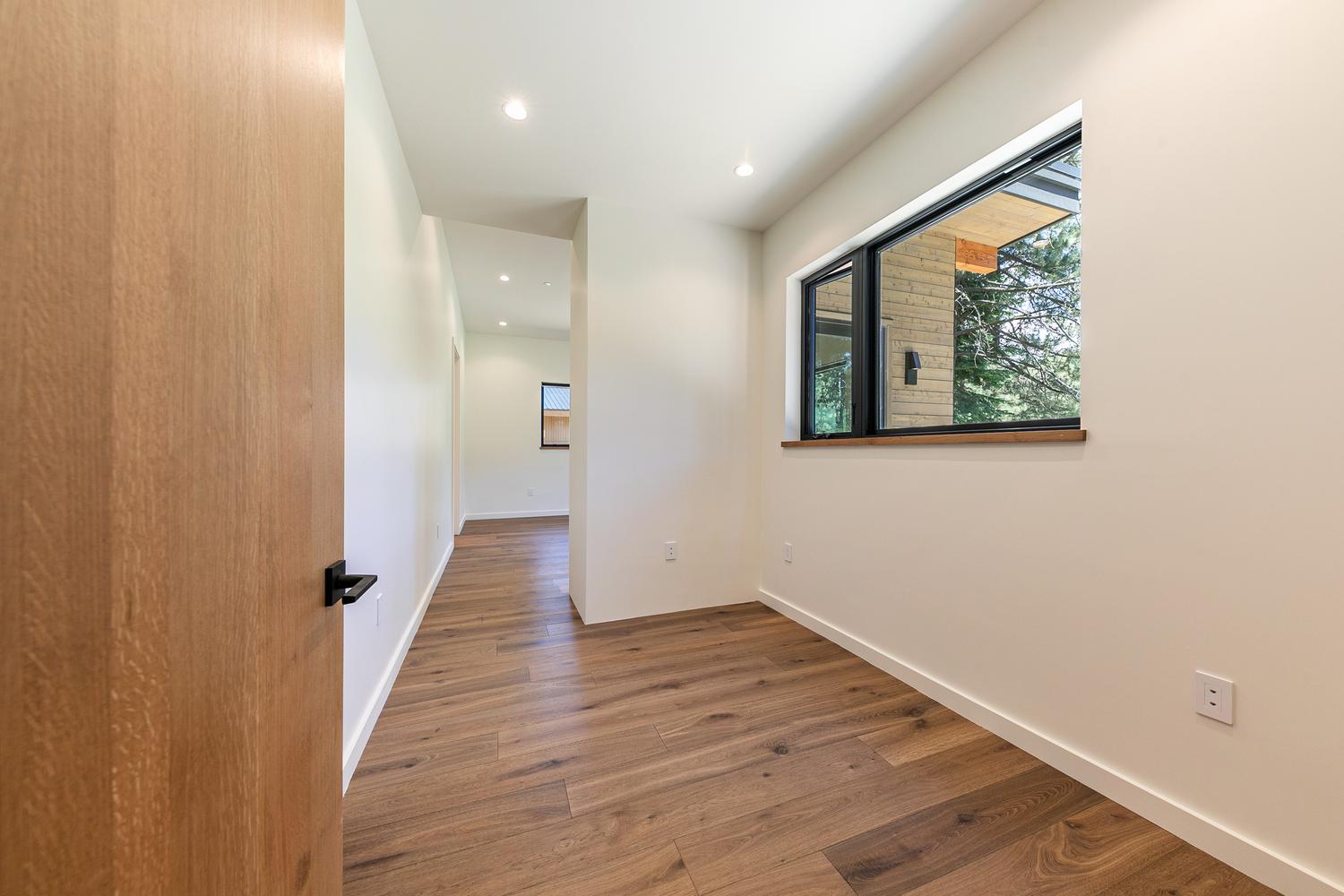 This interior view captures a bright, modern hallway leading into an adjacent room, showcasing a clean aesthetic with white walls and warm-toned wood-look flooring. A large window with a dark frame provides natural light and a view of the exterior, while a wooden door with a matte black handle stands in the foreground. The perspective draws the eye through the transition space, highlighting the minimalist design and open flow of the home.
