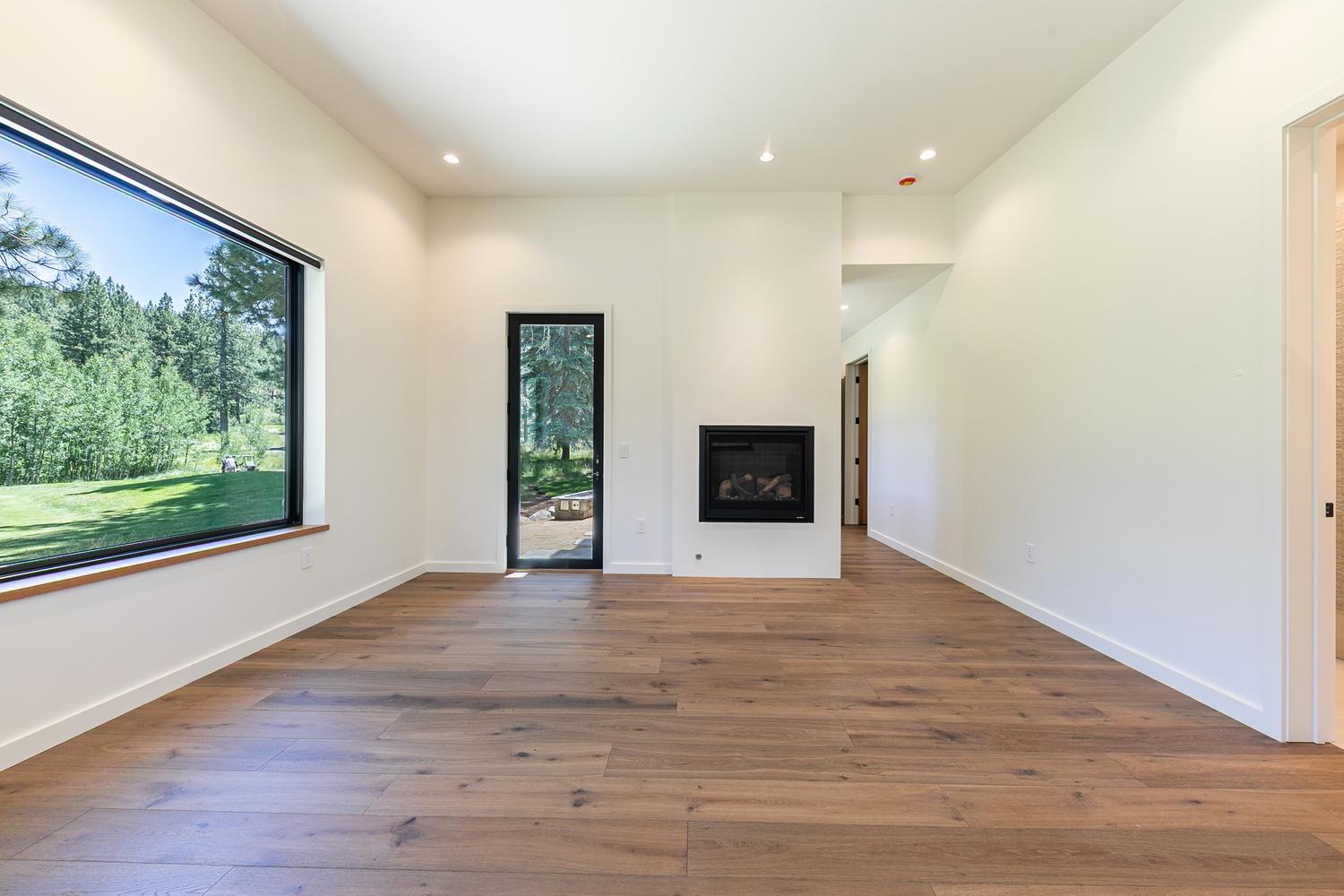 This bright and minimalist living room features wide-plank wood flooring and crisp white walls, creating an airy and modern atmosphere. A large window on the left provides a scenic view of the lush green outdoors, while a glass-paned door offers direct access to the exterior. A sleek, black-framed fireplace is centered on a prominent wall, serving as a clean focal point for the space.