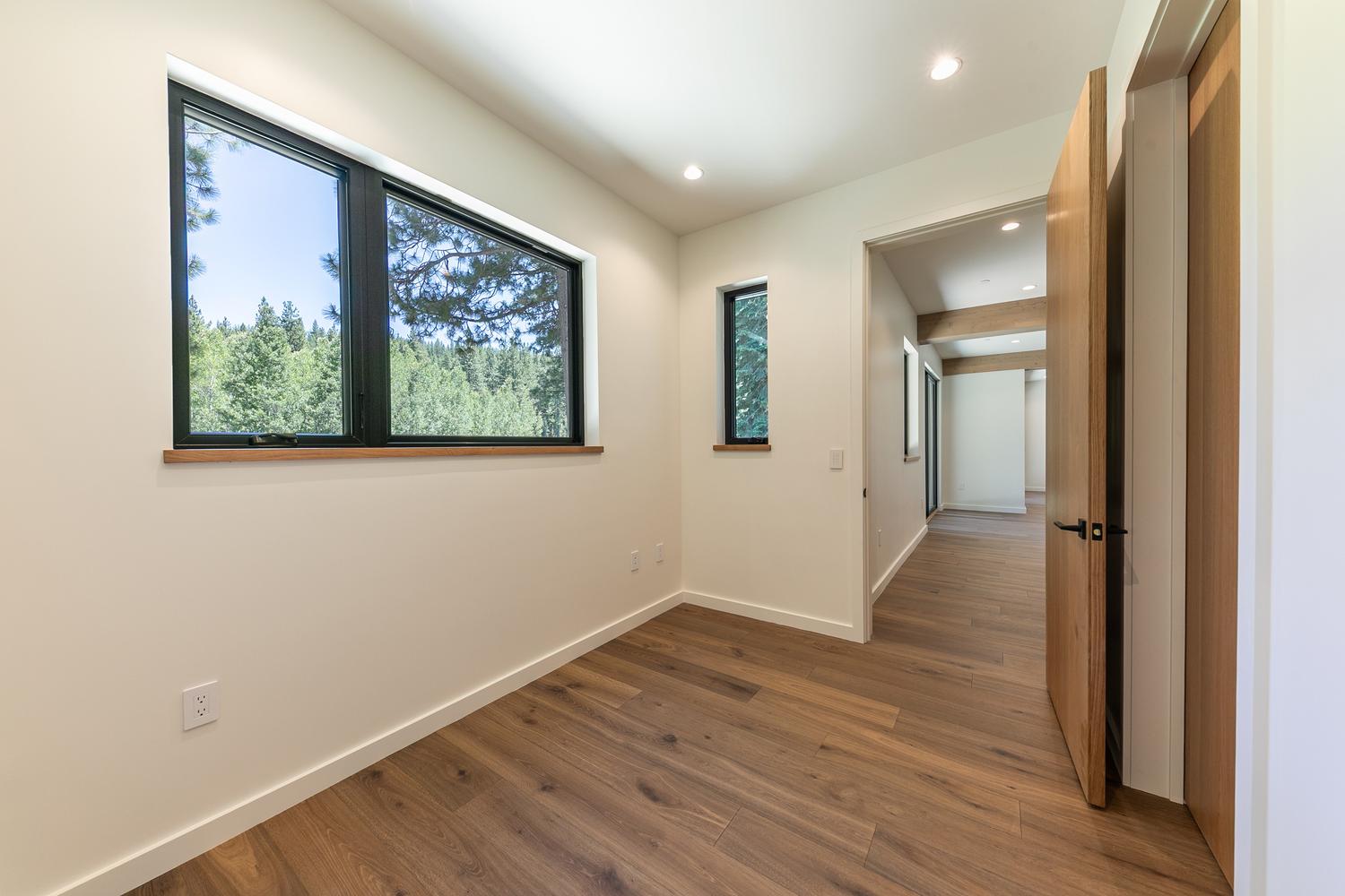 This bright, minimalist room features warm wood-tone flooring and crisp white walls, creating a clean and versatile space. A large window with a dark frame provides a scenic view of the surrounding forest, while a smaller adjacent window adds extra natural light. The room opens into a hallway, showcasing a modern wooden door and a seamless transition between living areas.