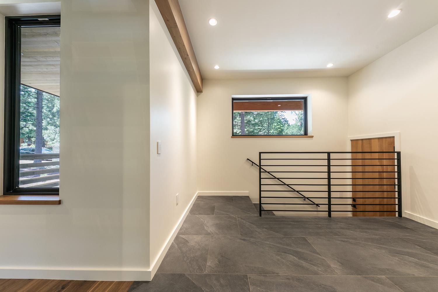 This modern landing area features dark, large-format tile flooring and crisp white walls, creating a clean and minimalist aesthetic. A black metal railing borders the staircase, while a horizontal window provides a view of the surrounding trees and natural light. The space is accented by a warm wood beam overhead and a wooden door, offering a balanced contrast between industrial elements and natural textures.