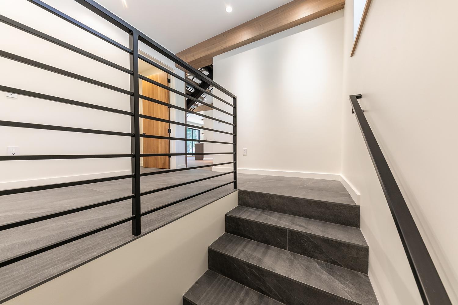 This modern staircase area features dark, large-format stone tiles on the steps and landing, contrasted by crisp white walls and a light wood ceiling beam. A sleek black metal railing with horizontal bars provides a contemporary safety barrier, while a matching black handrail is mounted to the wall. The perspective looks up the stairs toward a landing that leads to a doorway with a light wood door, creating a clean, minimalist aesthetic.