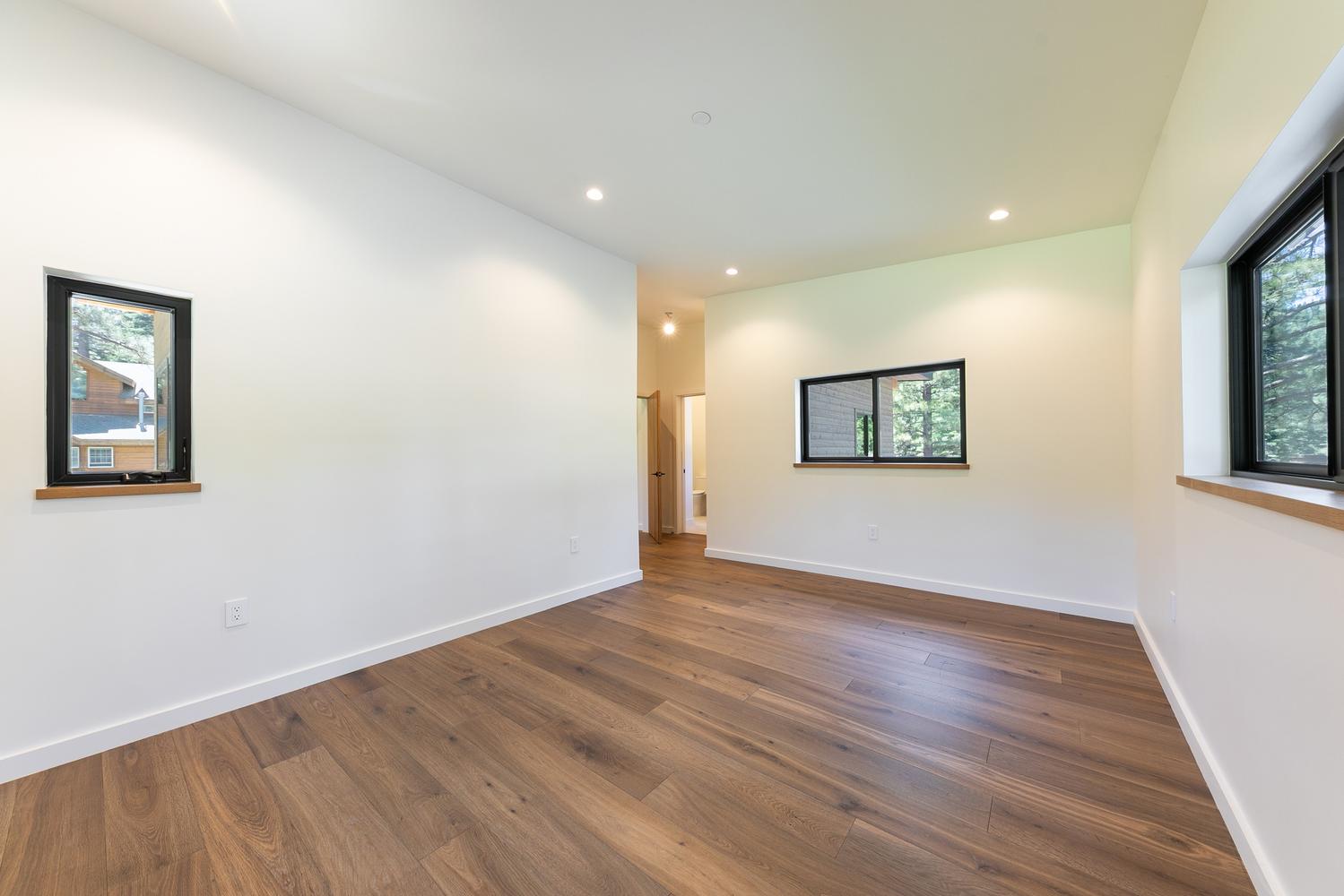 This bright, minimalist bedroom features warm-toned wood flooring and crisp white walls, creating a clean and inviting atmosphere. The room is illuminated by recessed ceiling lights and natural light streaming through three black-framed windows of varying sizes. A doorway in the background leads to an adjacent hallway, highlighting the open and airy layout of the home.