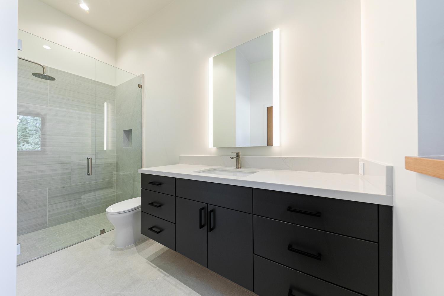 This modern bathroom features a sleek, floating vanity with dark cabinetry and a light-toned stone countertop. A large, rectangular mirror with integrated side lighting hangs above the sink, while a glass-enclosed walk-in shower with light-colored tile and a rainfall showerhead occupies the corner. The space is finished with neutral floor tiles and clean, minimalist lines, creating a bright and contemporary atmosphere.