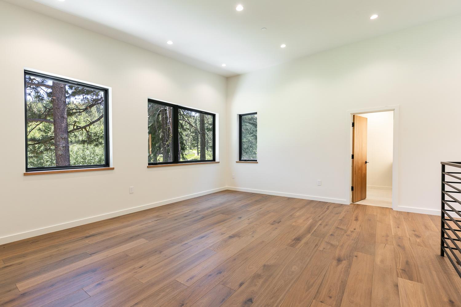 This bright, minimalist room features wide-plank wood flooring and crisp white walls, creating an airy and open atmosphere. Three large windows with black frames provide ample natural light and views of the surrounding forest, while a wooden door leads to an adjacent space. The room's clean lines and neutral palette offer a versatile canvas for any interior design style.