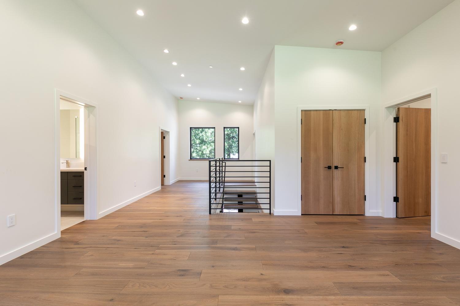 This bright, modern landing area features wide-plank wood flooring and crisp white walls, creating an airy and open atmosphere. A central staircase with a sleek black metal railing serves as a focal point, leading down to a lower level, while double light-wood doors and a doorway leading to a bathroom provide access to adjacent rooms. The space is illuminated by numerous recessed ceiling lights, emphasizing the clean lines and contemporary architectural design.
