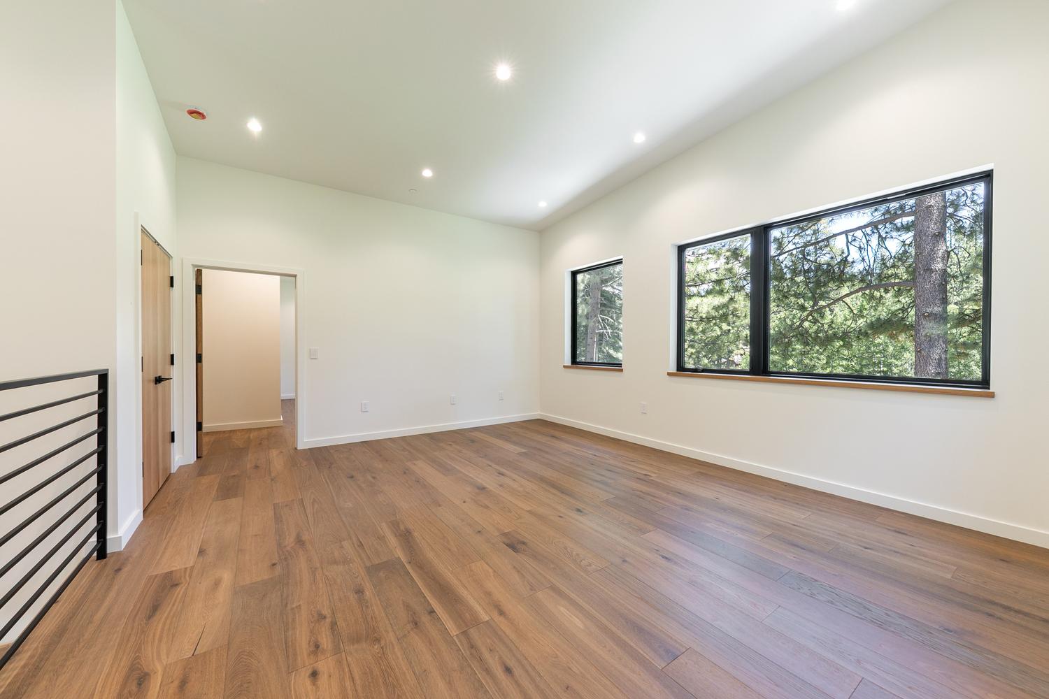 This bright, minimalist room features light-toned wood flooring and crisp white walls, creating an airy and versatile living space. Large windows with black frames provide a clear view of the surrounding trees, while recessed lighting illuminates the room from above. A wooden door leads to an adjacent hallway, and a black metal railing is visible on the left, suggesting an open-concept layout.