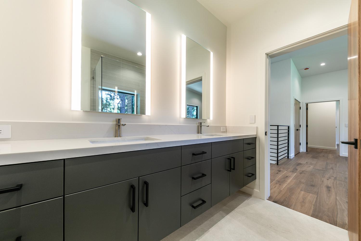 This modern primary bathroom features a sleek, floating double vanity with dark gray cabinetry and a crisp white countertop. Two rectangular mirrors with integrated vertical lighting are mounted above the sinks, creating a clean and contemporary aesthetic. The view extends through an open doorway into a hallway with wood flooring and a metal staircase railing, emphasizing the home's open and airy layout.