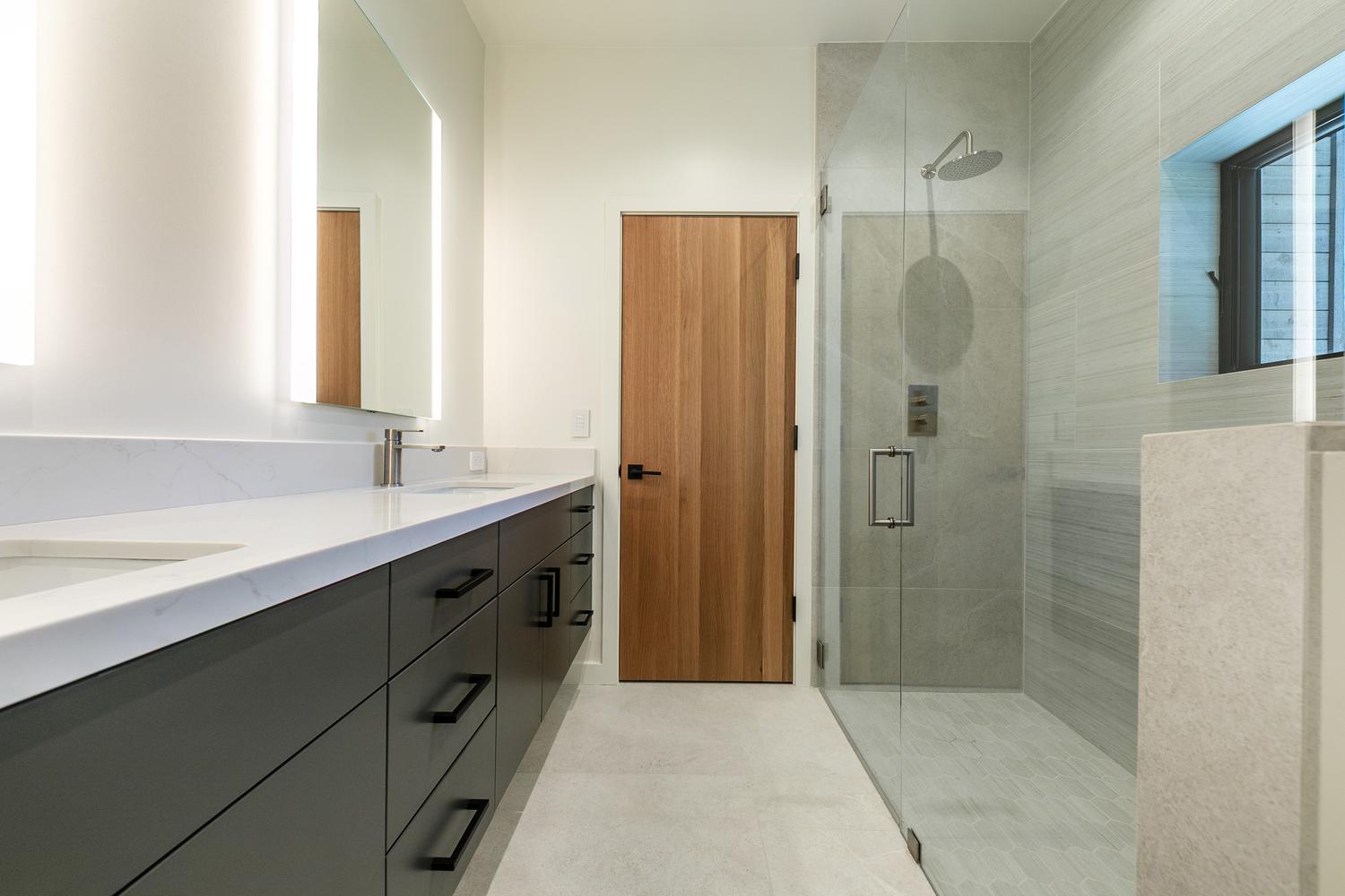 This modern bathroom features a long vanity with dark cabinetry, a white countertop, and a large, illuminated mirror. To the right, a glass-enclosed walk-in shower showcases textured wall tiles and a rainfall showerhead. A light wood door adds warmth to the clean, neutral aesthetic of the space.