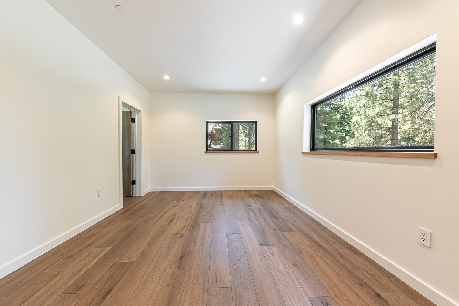 This bright, minimalist room features light-colored walls and warm-toned wood flooring, creating a clean and versatile living space. The room is illuminated by recessed lighting and two windows, including a large horizontal window that provides a scenic view of the surrounding trees. A doorway on the left leads to an adjacent area, and the overall aesthetic is modern and neutral, making it suitable for a bedroom or home office.
