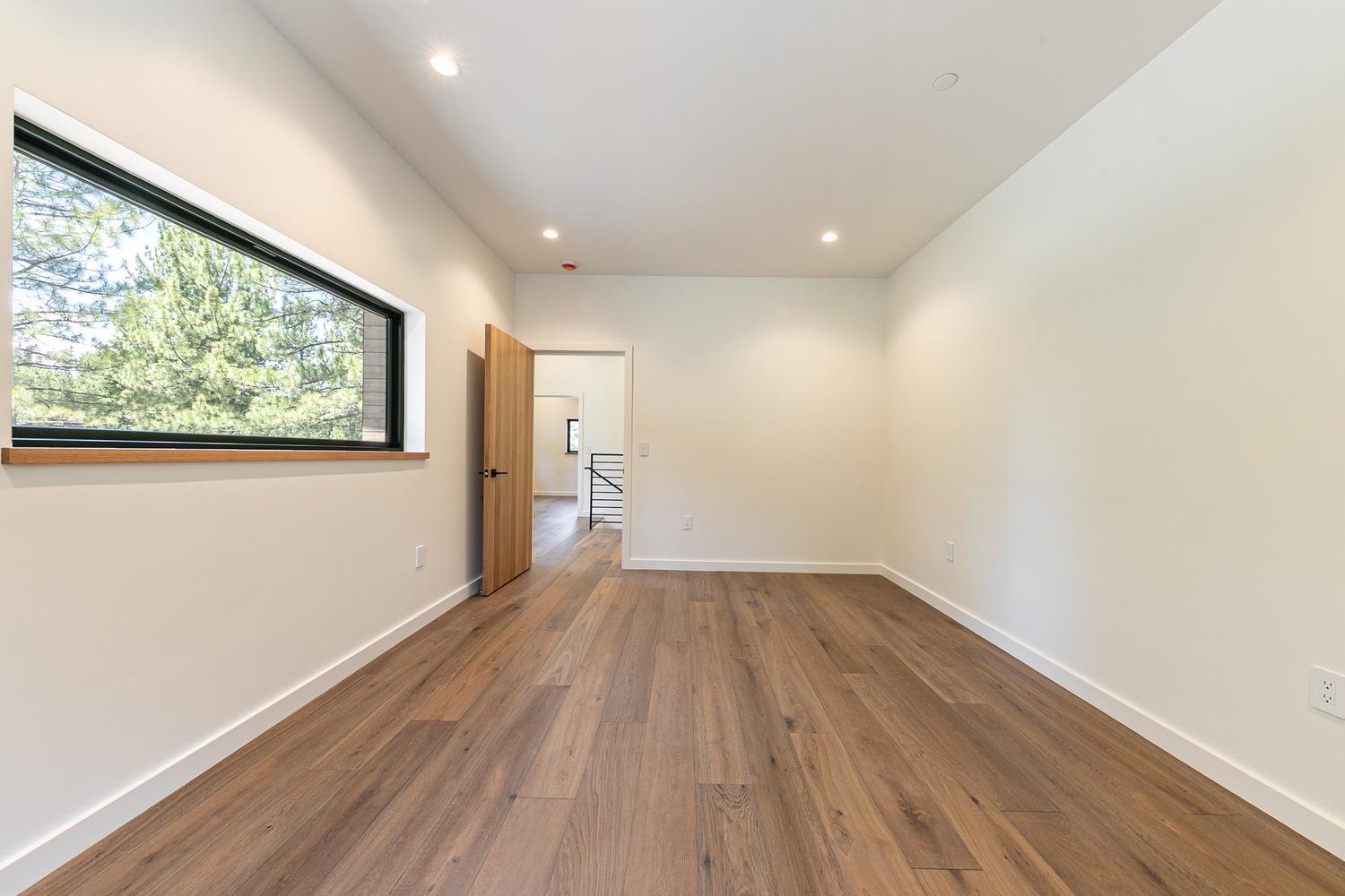 This bright, minimalist room features light-toned walls and warm-toned wood-look flooring, creating an airy and versatile living space. A wide, horizontal window with a dark frame provides a view of the surrounding trees, while a wooden door leads to an adjacent hallway. The perspective is centered, highlighting the clean lines and neutral aesthetic of the room.
