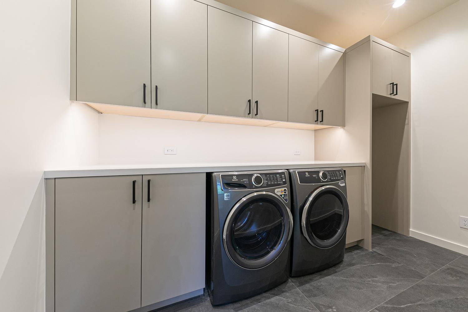 This modern laundry room features sleek, light-toned cabinetry with dark hardware, providing ample storage space above and beside a pair of dark-finished front-load washer and dryer units. A clean, white countertop spans the base cabinets, offering a functional workspace, while under-cabinet lighting illuminates the area. The room is finished with dark, large-format floor tiles, creating a sophisticated and organized utility space.