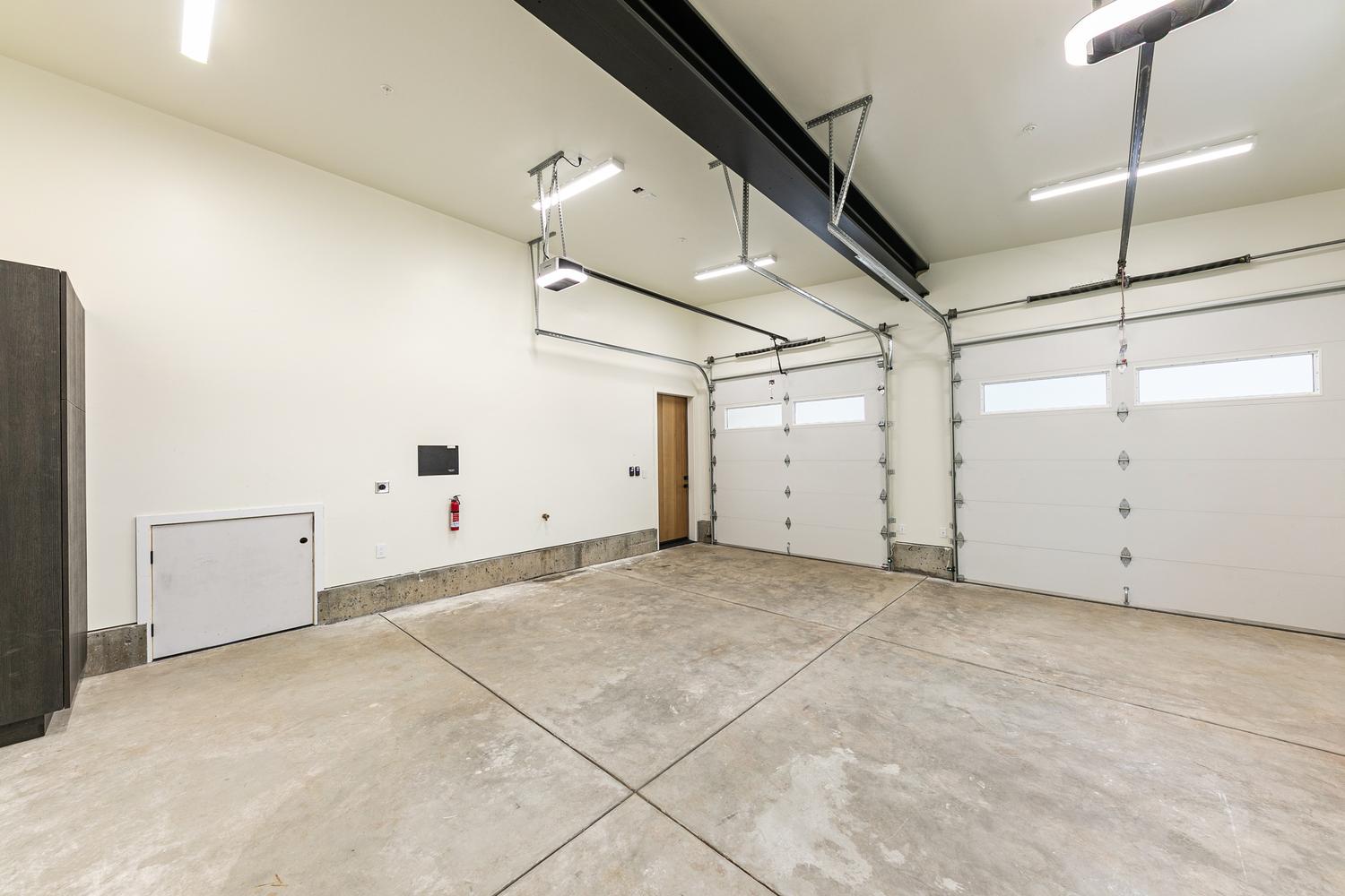 This spacious, clean-lined garage features two white sectional doors with windows, providing ample room for multiple vehicles. The space is finished with smooth, light-colored walls, a concrete floor, and exposed structural steel beams overhead. A small wooden door leads to an interior area, and a fire extinguisher is mounted on the wall, emphasizing safety and utility in this functional storage space.