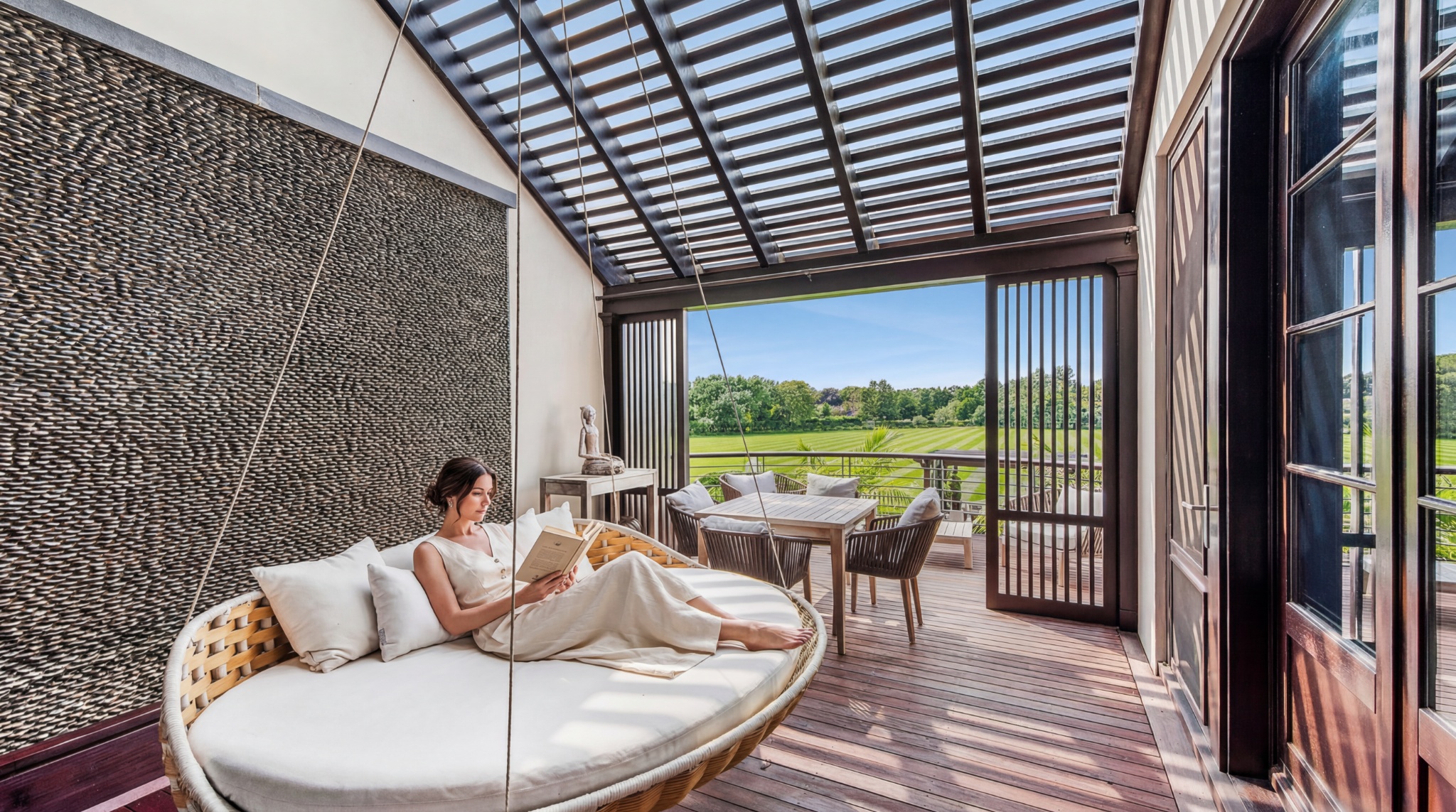 This serene, covered balcony features a high-end hanging daybed with white cushions, providing an ideal spot for relaxation. The space showcases high-end materials, including a textured stone feature wall and wooden decking, seamlessly transitioning to an open-air dining area overlooking a manicured lawn. The design exudes luxury and tranquility, perfectly blending indoor comfort with expansive outdoor scenery.