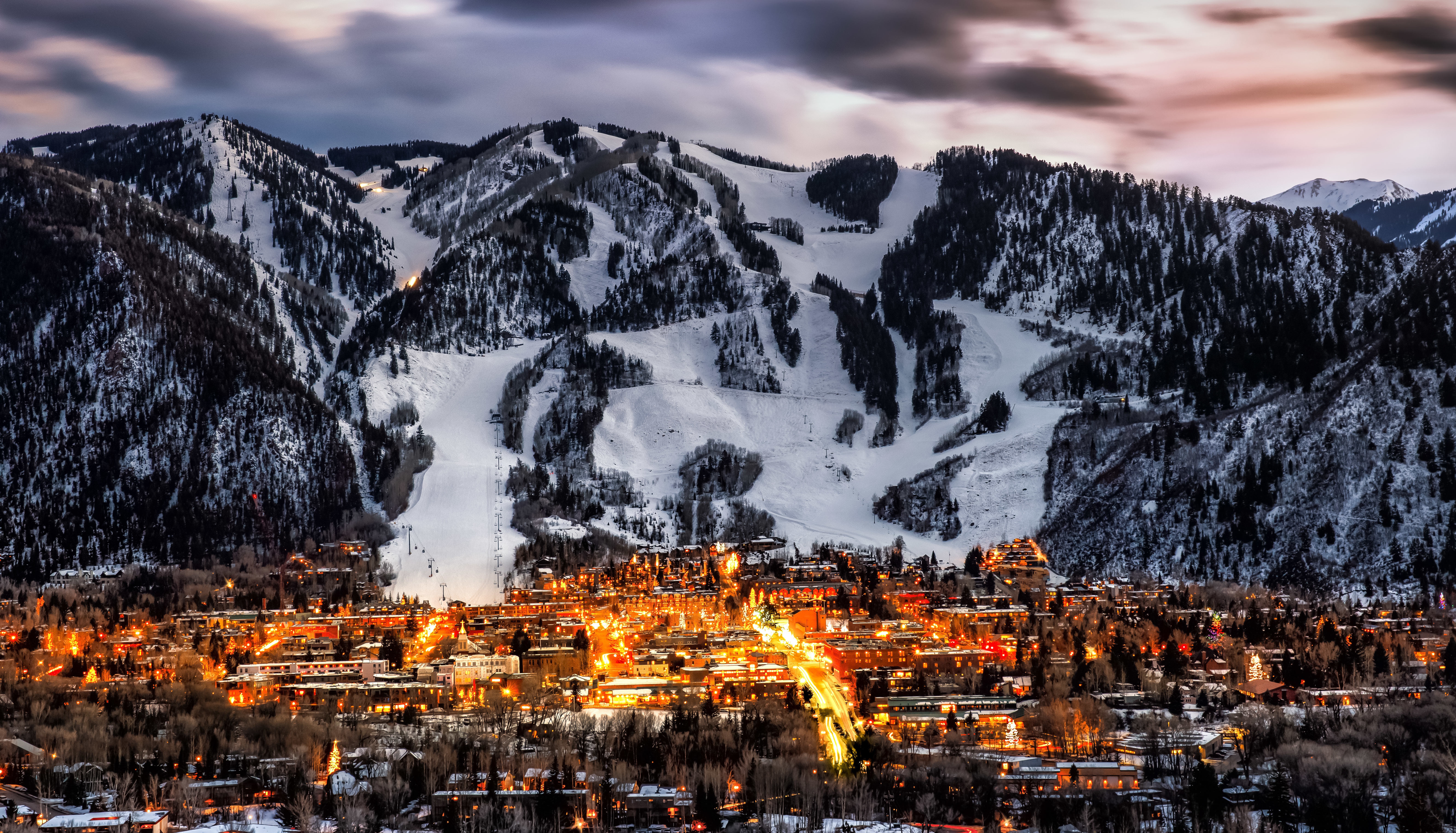 This aerial view showcases a luxurious property nestled in a mountain setting. The town is illuminated with warm lights, contrasting beautifully with the snow-covered slopes. The image highlights the proximity to ski resorts and the overall picturesque location, perfect for buyers seeking a scenic retreat.