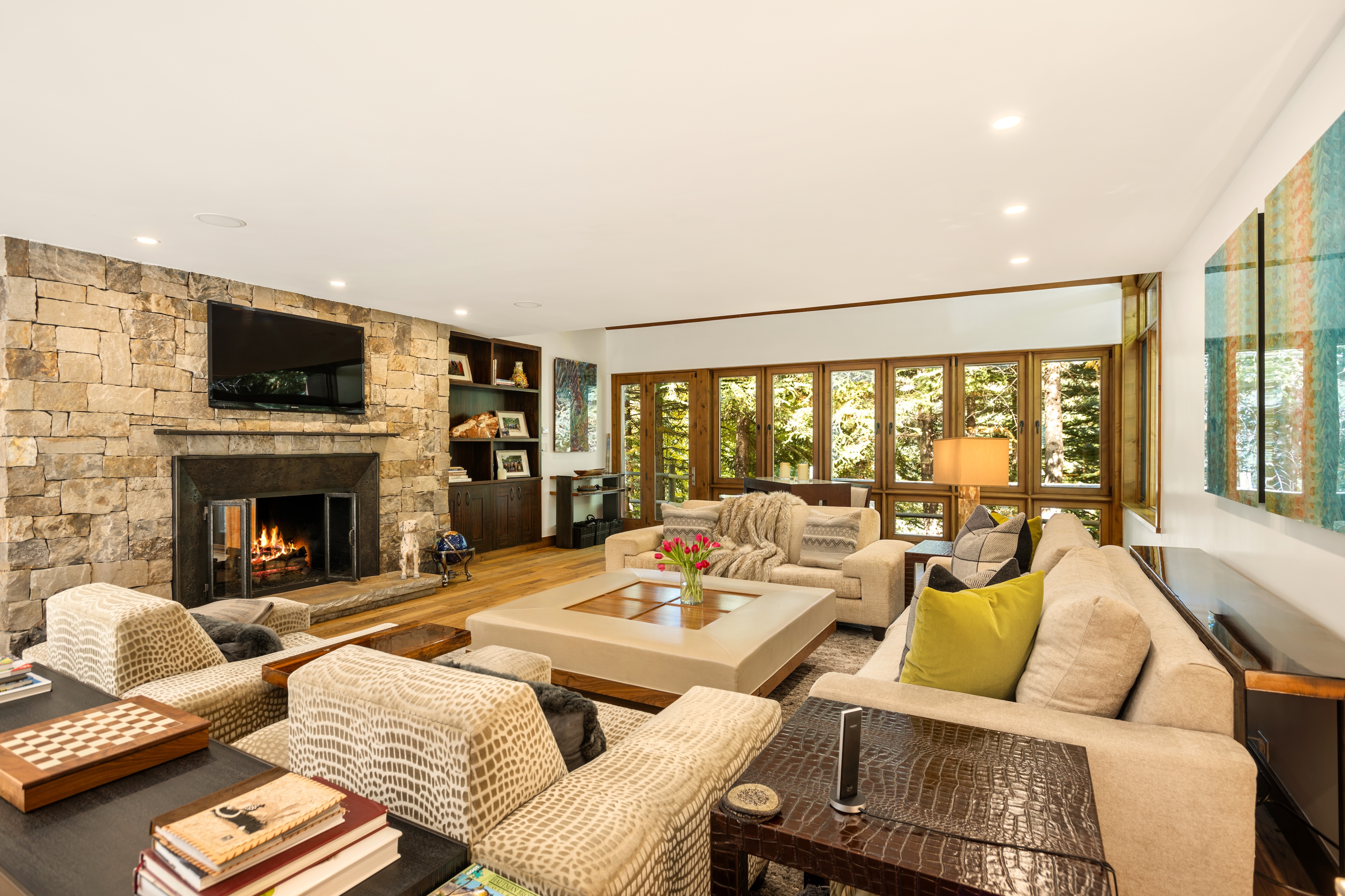 This is a warm and inviting living room, highlighted by a stone fireplace with a mounted TV above. The room features comfortable seating arrangements, including sofas and armchairs, arranged around a large, light-colored coffee table. Natural light floods the space through the many windows, creating a bright and appealing atmosphere.