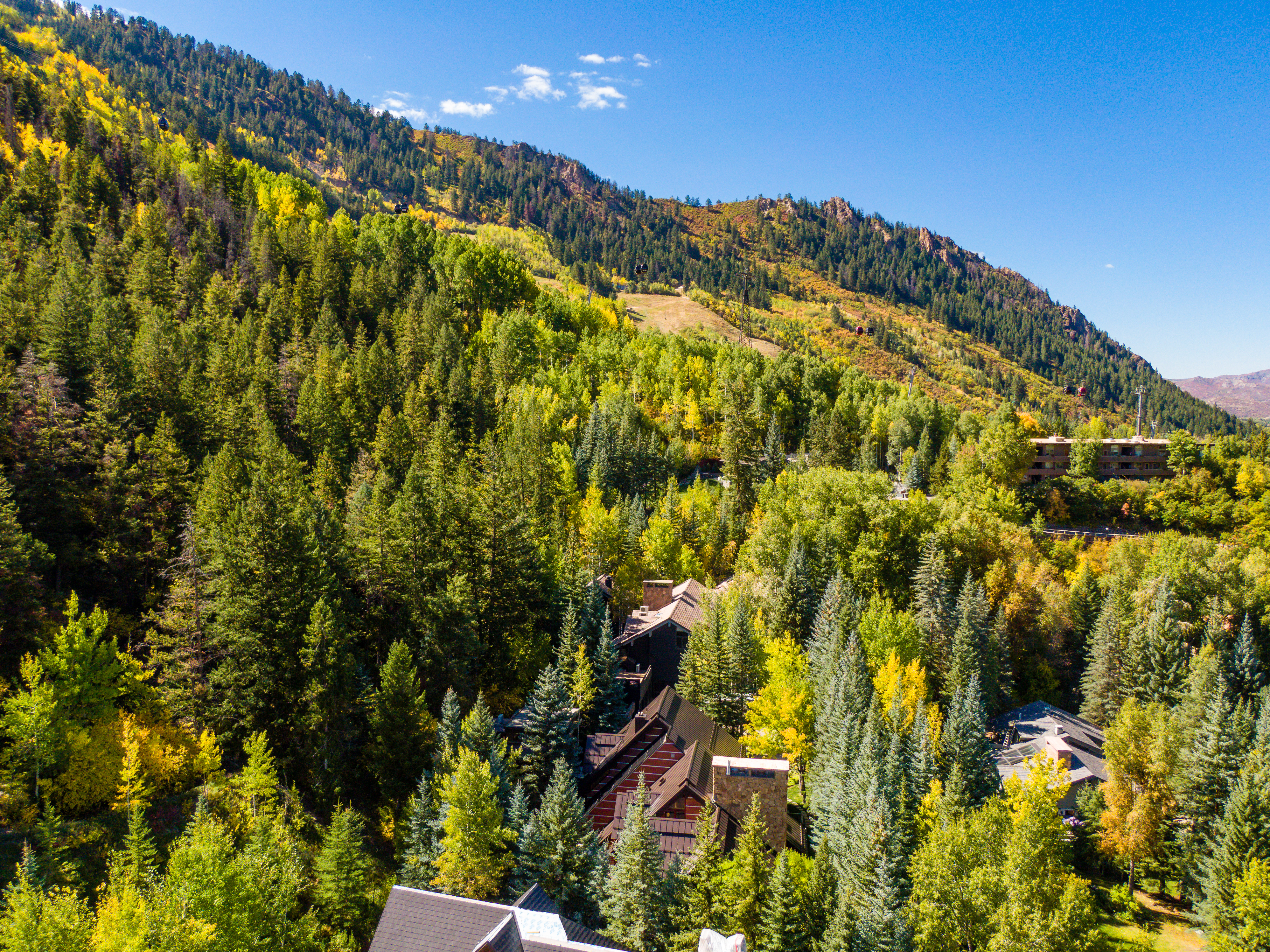 This aerial view showcases a mountain home nestled amongst lush greenery with the surrounding mountains in the backdrop. The architecture features varying rooflines and natural materials that appear seamlessly integrated into the landscape. The image highlights the property's secluded and scenic location, which is a desirable feature for buyers seeking privacy and natural beauty.