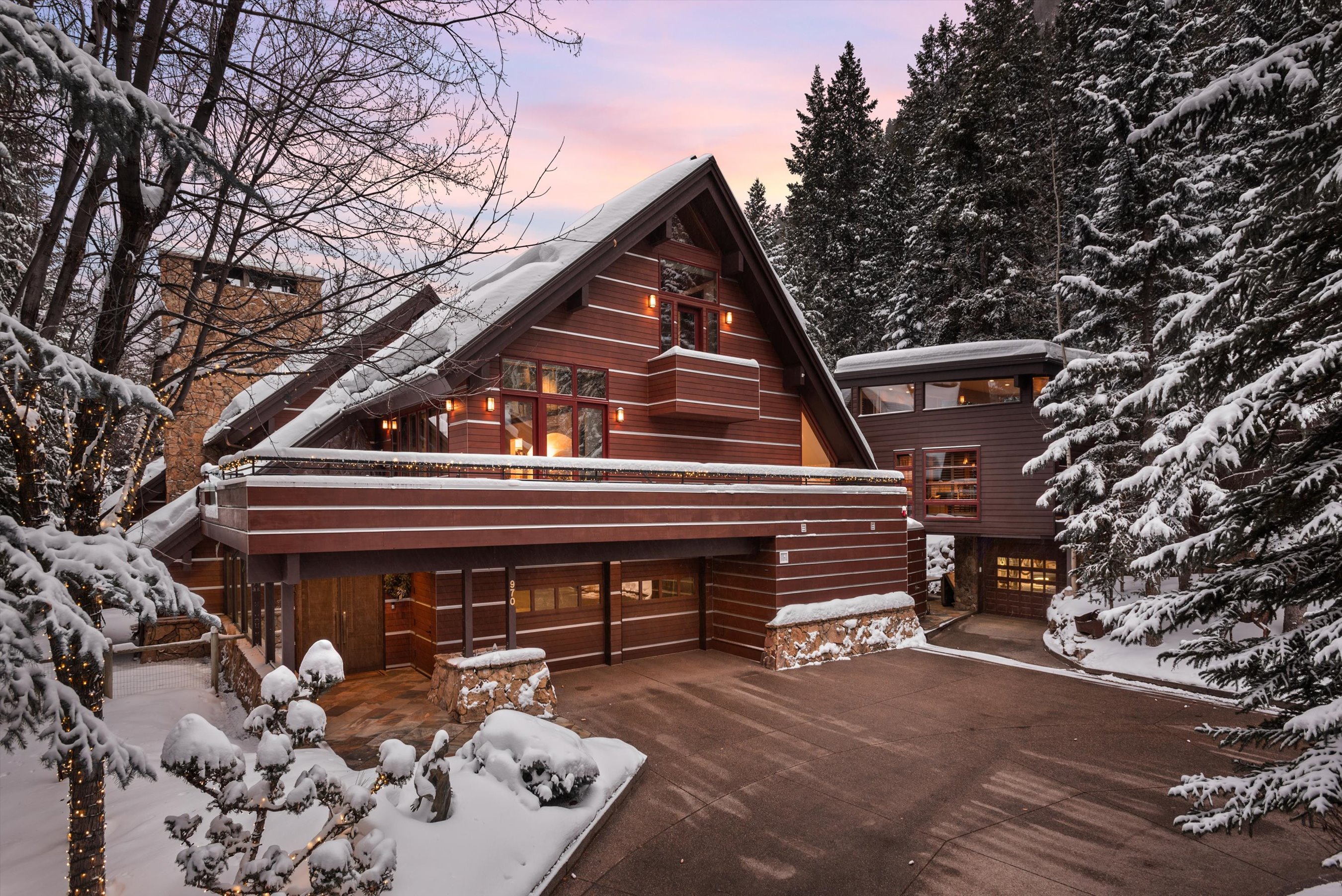 This is a front view of a luxurious, chalet-style home in a snowy, wooded setting. The exterior features a mix of wood siding and stone accents accentuating its architectural design. The house is well-lit, adding to its inviting appearance, with a spacious driveway leading up to a multi-car garage.